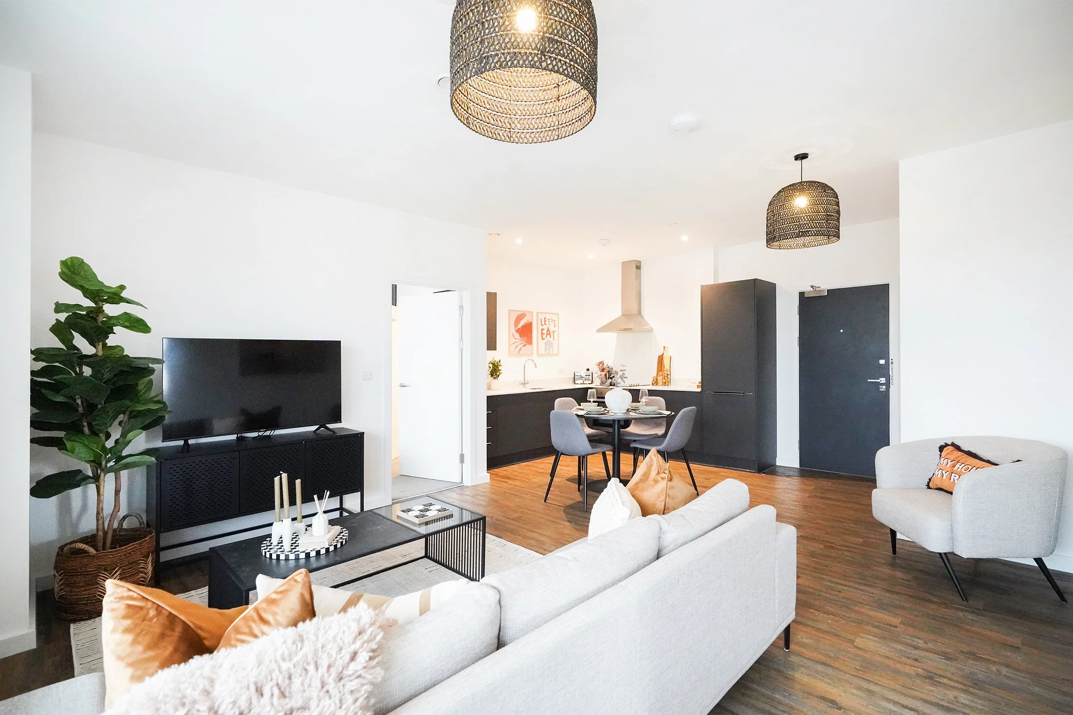A modern open-concept living room and kitchen area with white walls, hardwood floors, two black woven pendant lights, a large flat-screen TV on a black media console, a white sofa with orange and white pillows, a black coffee table with candles, a potted fig plant, a round dining table with four gray chairs, a black refrigerator, black cabinetry, and artwork on the wall near the kitchen.