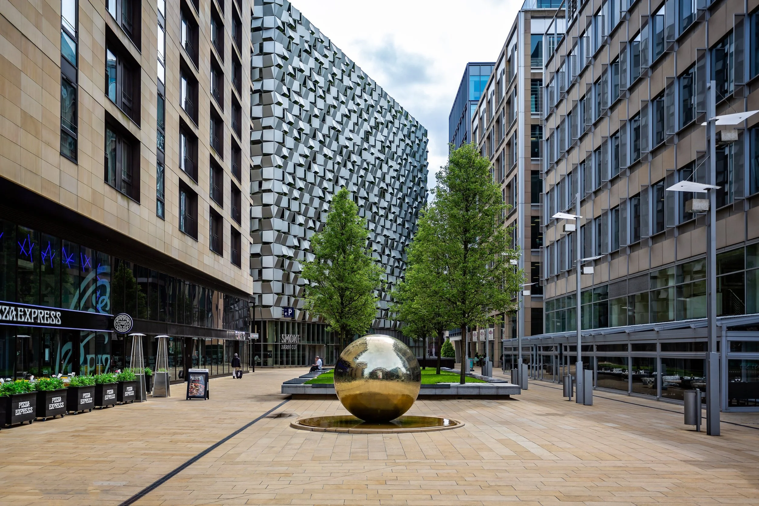 City plaza with modern glass and stone buildings, trees, a large reflective golden sphere fountain, and outdoor seating areas.