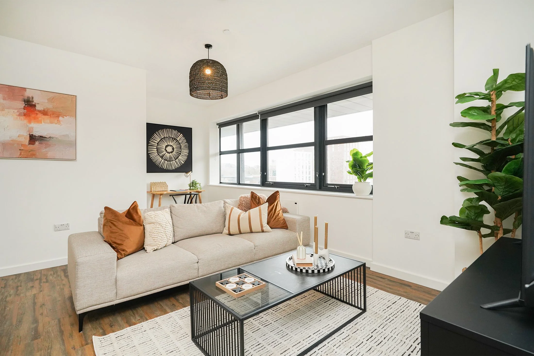 A modern living room with a beige sofa adorned with brown and cream pillows, a black coffee table with a decorative tray, large windows with dark frames, and artwork with abstract and circular designs on the walls. There is a leafy green plant to the right and a black woven pendant light hanging from the ceiling.