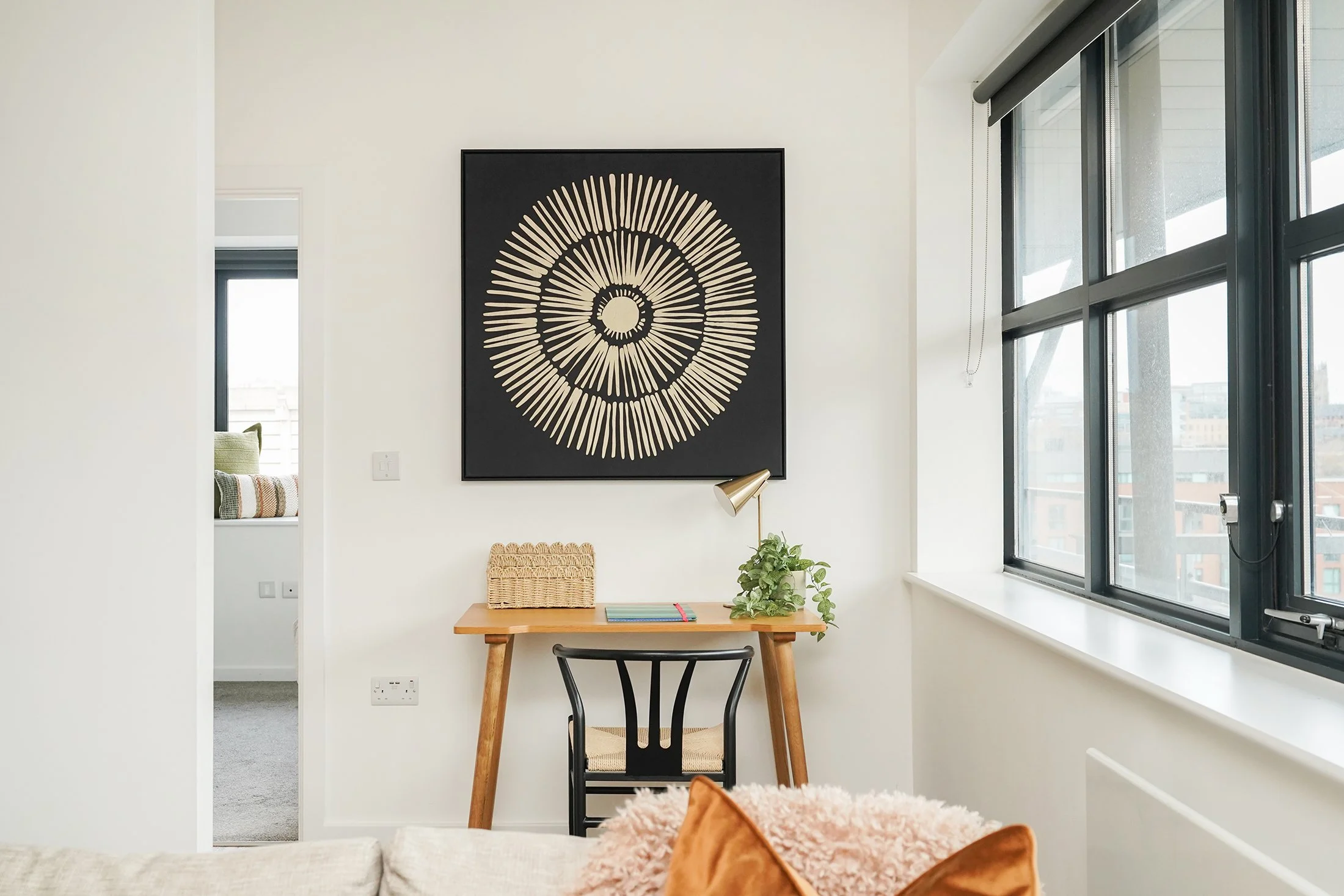 Interior of a modern apartment with a black and beige radial art piece on the wall, a wooden table with a green plant and notebooks, a black chair, and large windows with city views.