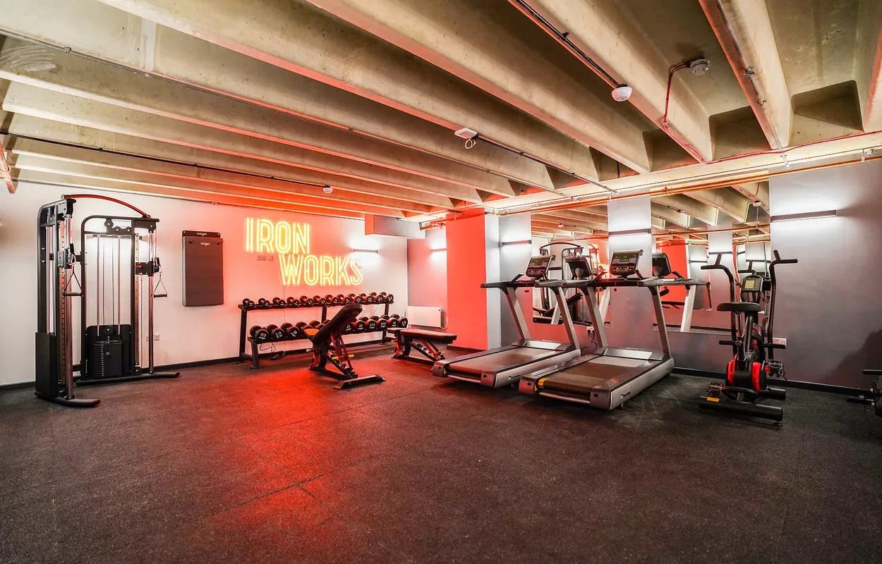 The modern gym at Iron Yard with cardio machines including treadmills and an exercise bike, weight equipment, and a neon sign that says and a rack of dumbbells. The space has an industrial ceiling and motivational lighting.