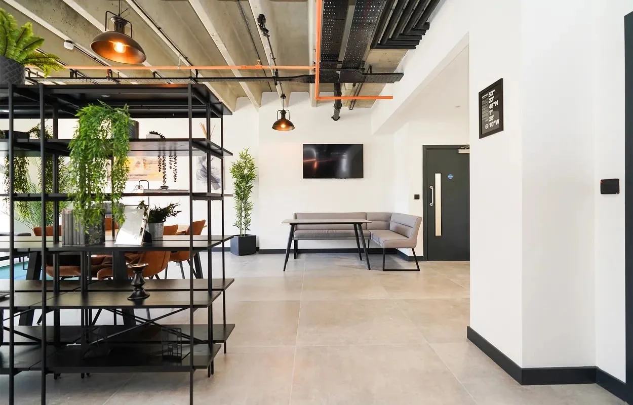Modern industrial-style interior with a dining area and seating nook, featuring a black metal shelving unit with potted plants, a high chair, and minimalistic furniture, exposed pipes and ducts on the ceiling, concrete and white walls, a wall-mounted TV, and a digital wall clock.