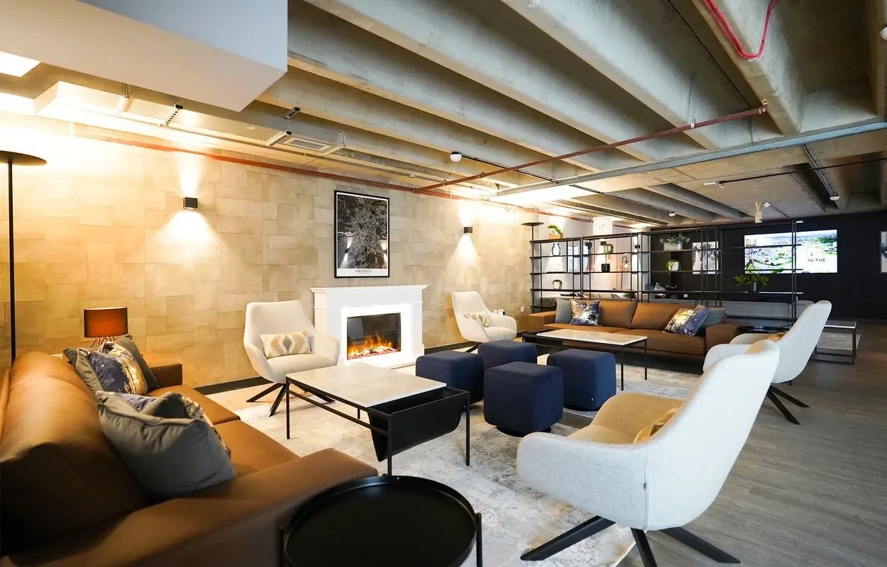 Modern lounge with beige and brown sofas, white armchairs, and blue ottomans, featuring a fireplace on a beige brick wall, decorative pillows, a black-and-white framed artwork, wall sconces, and a black room divider with shelving, illuminated by warm lighting.
