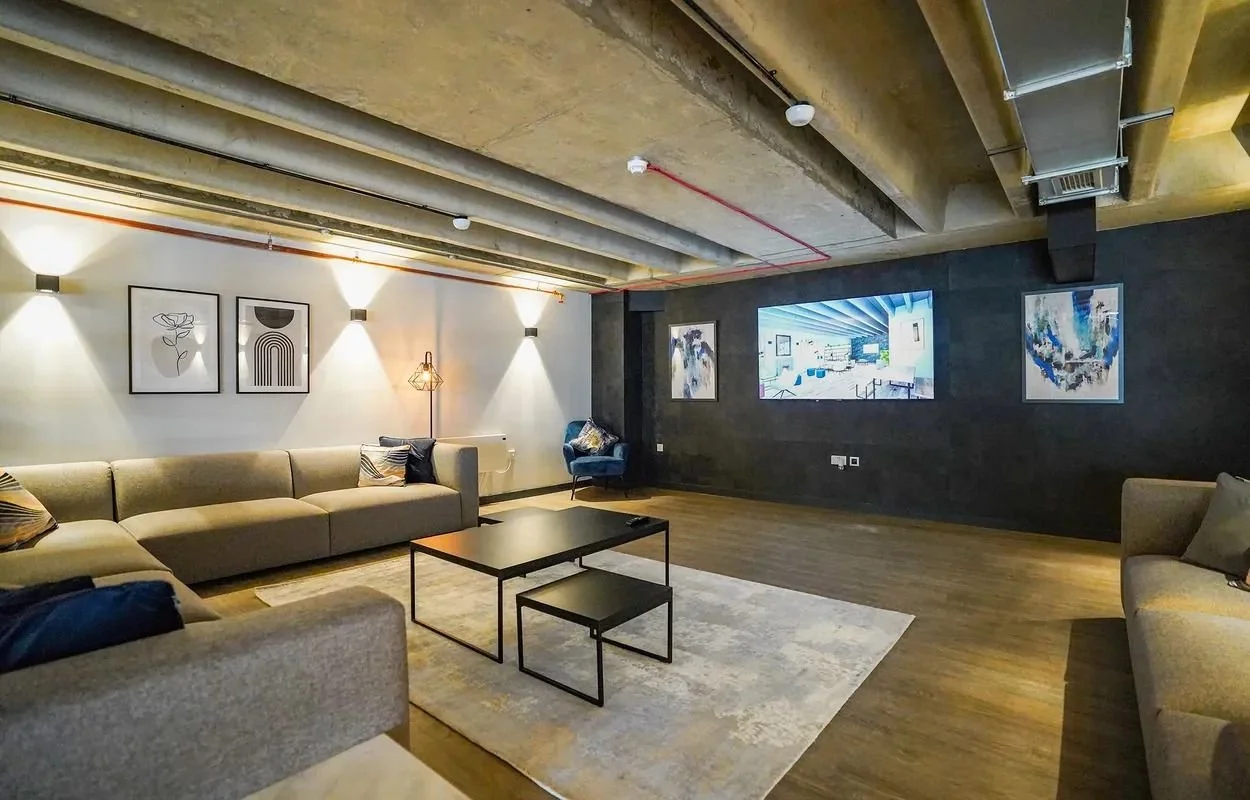 Modern living room with beige sofas, black coffee table, patterned pillows, abstract artwork on the wall, and a large television on a dark wall.