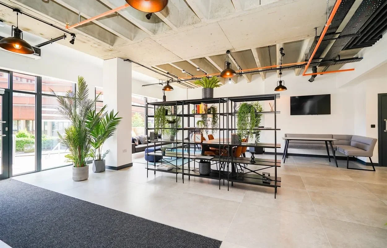 Modern office lobby with large windows, potted plants, a black metal shelving unit with various decorations, a sitting area with chairs and a television, exposed concrete ceiling with pendant lights, and a combination of carpet and tile flooring.