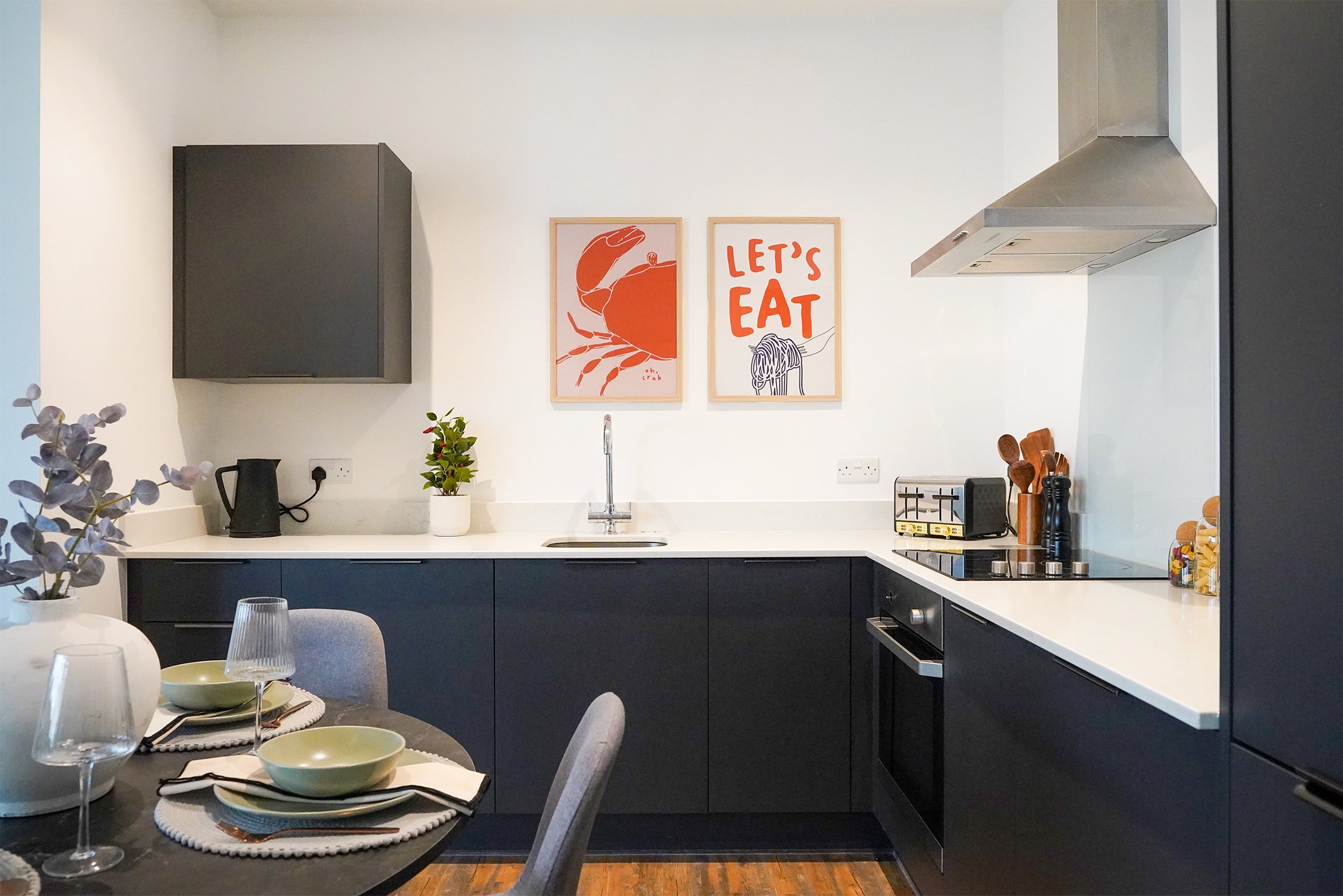 A modern kitchen with dark lower cabinets, a white countertop, and a stainless steel range hood. Decor includes two framed art prints on the wall, one depicting a crab, and the other with the phrase 'Let's Eat' with a drawing of a cat. The dining tab