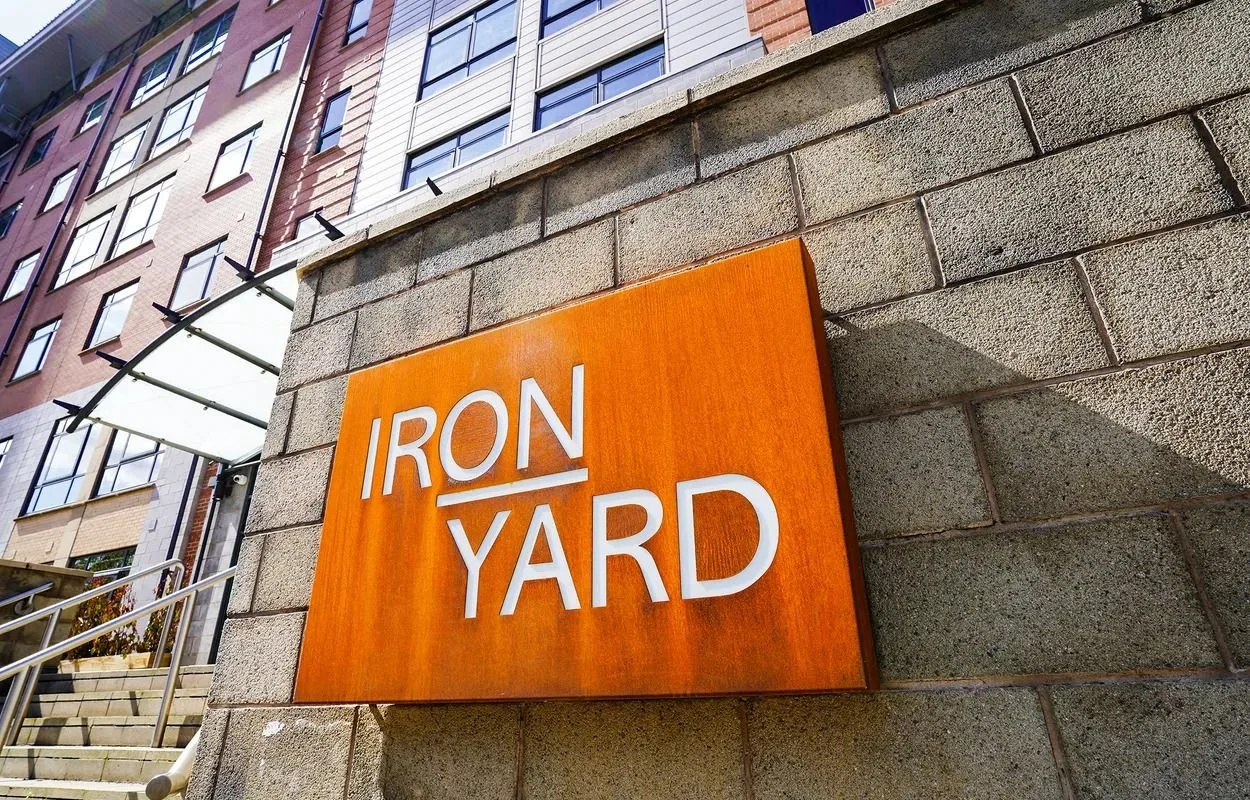 Sign on a brick wall that reads "IRON YARD" in white letters on an orange background, with apartment buildings and stairs in the background.