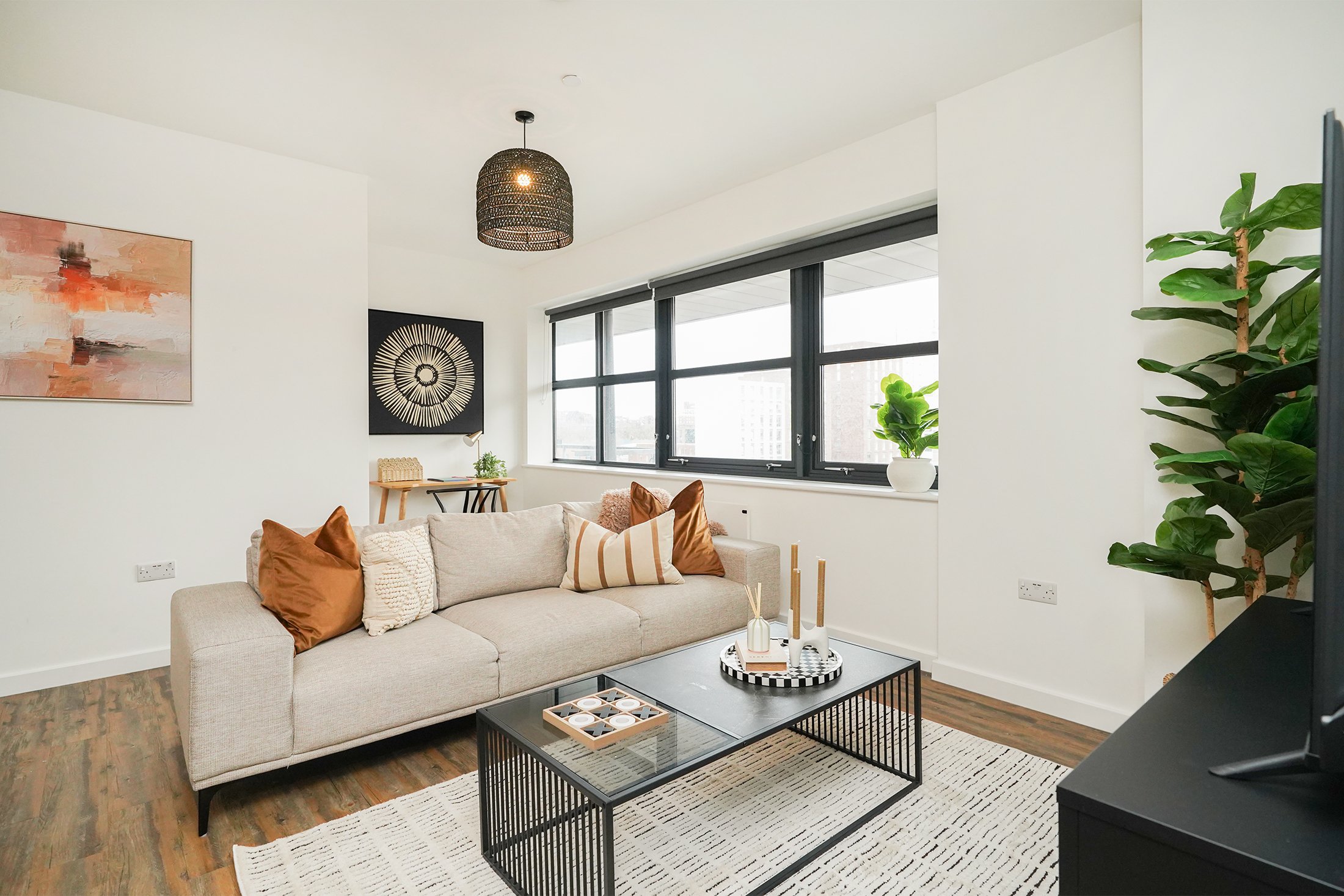 Modern living room with a beige sofa, brown pillows, and a black coffee table on a white striped rug, large window with roller blinds, abstract wall art, and a tall green plant in a white pot.