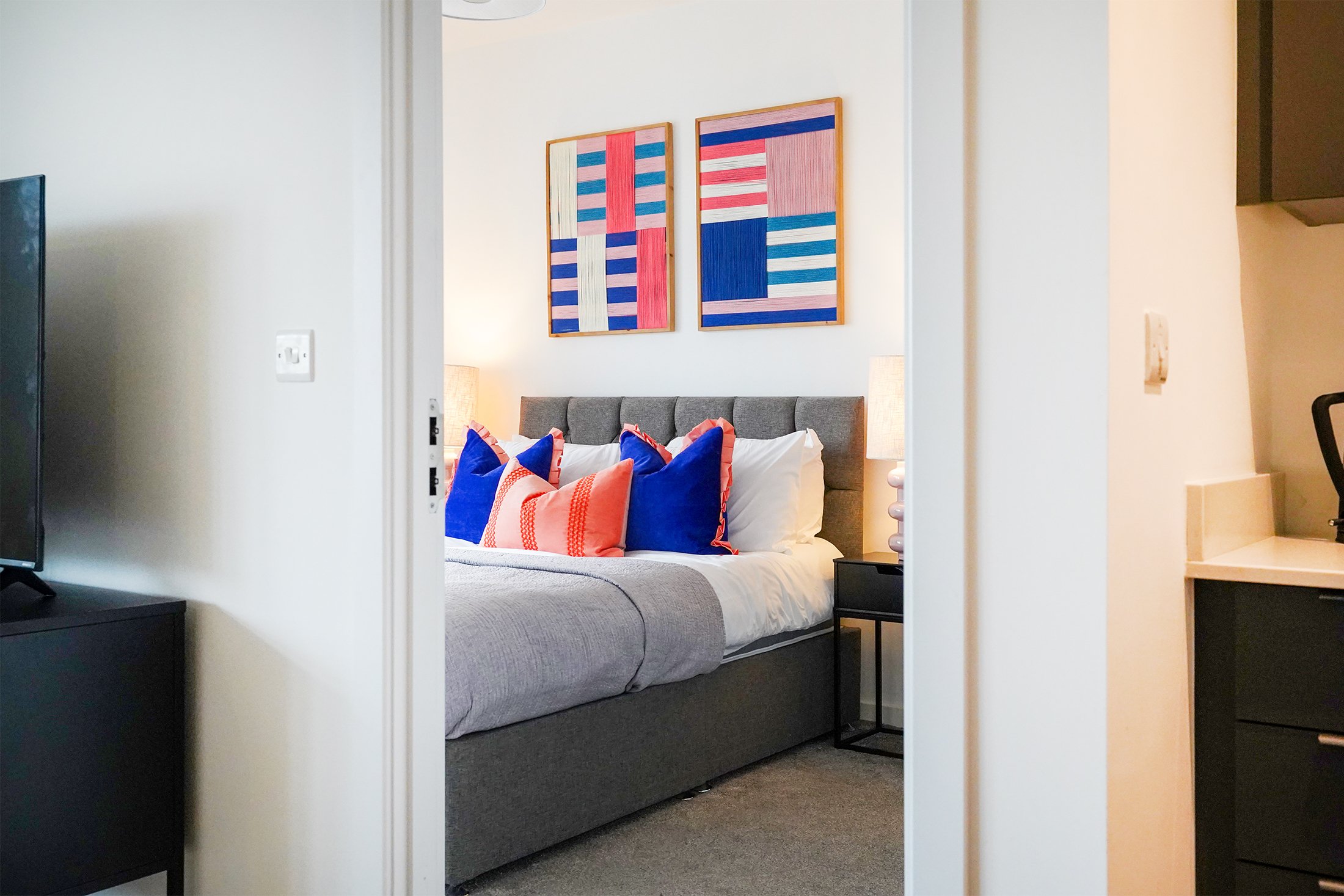 A bedroom with a gray upholstered bed, decorated with colorful blue and orange pillows, a light wood headboard, and abstract artwork with geometric patterns hanging on the wall.