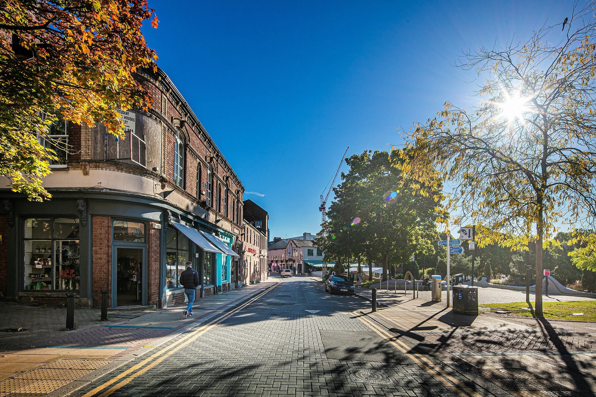 A Sheffield street scene with brick buildings on the left, trees with fall foliage on both sides, and a few pedestrians walking. The sky is clear and sunny, with the sun shining brightly.