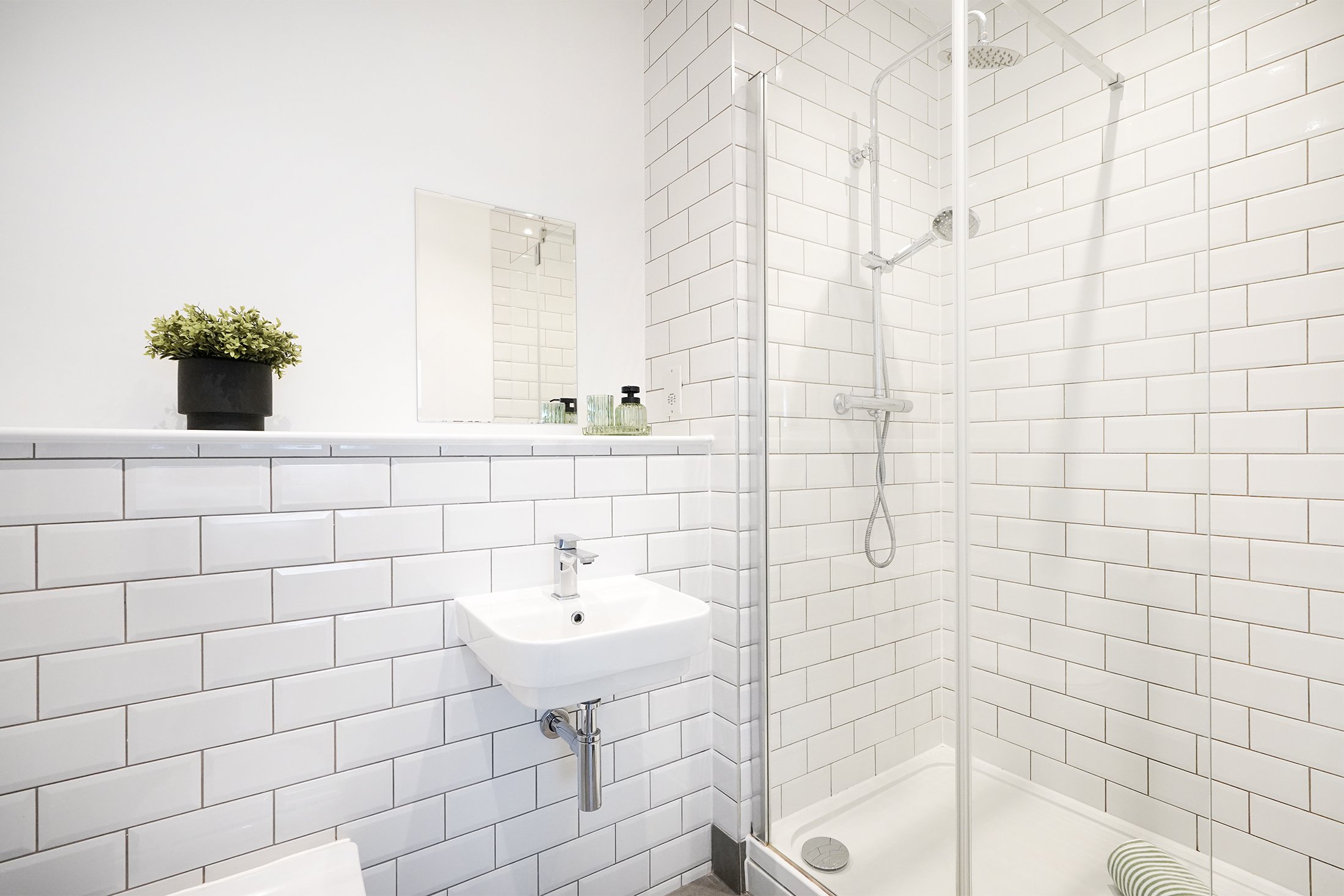 A modern bathroom with white subway tile walls, a wall-mounted sink with a chrome faucet, a mirror, a black potted plant, and a walk-in glass shower with a rainfall showerhead and a hand-held shower.