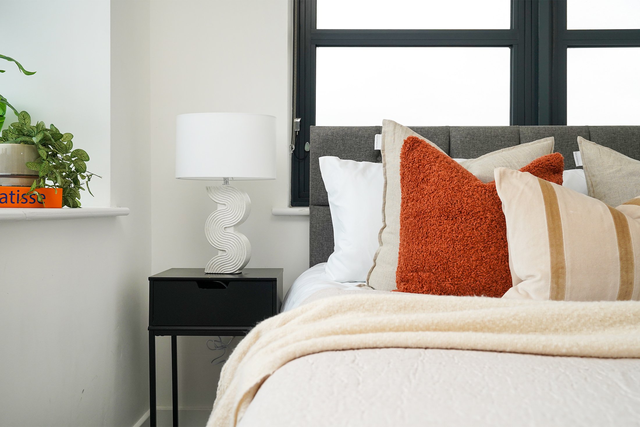 A bedroom with a bed featuring multiple pillows, including a textured orange pillow, a beige pillow, and a white pillow. There is a black nightstand with a white, curvy table lamp and a window with black framing behind the bed. A potted plant sits on a white shelf to the left, partially visible.