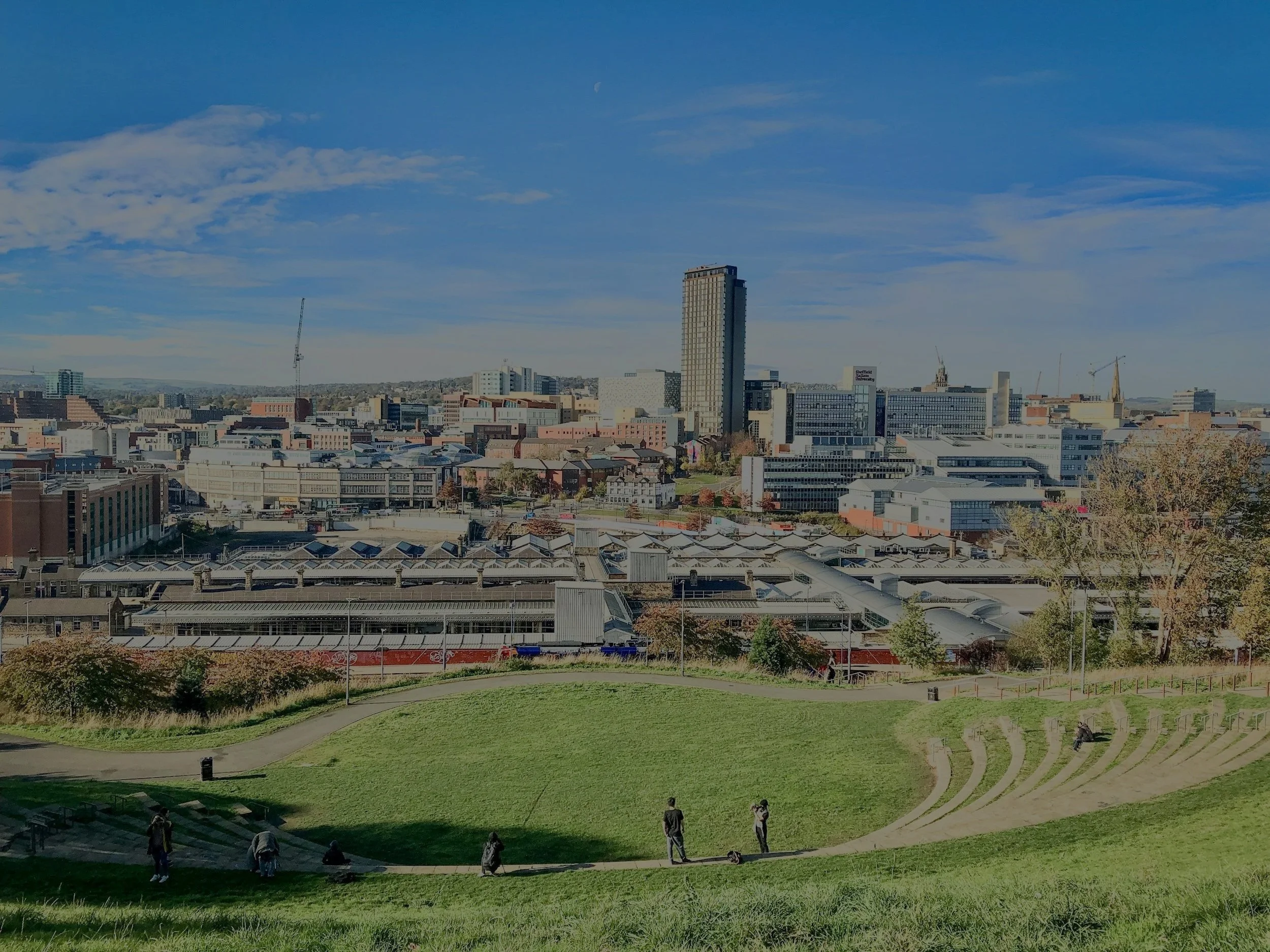 A Sheffield city skyline with tall buildings under a partly cloudy sky, viewed from a grassy park with walking paths and people sitting and walking.
