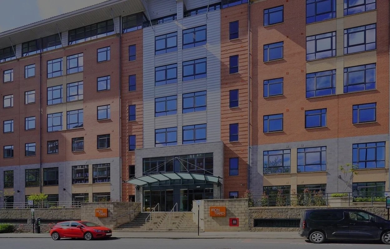 Front view Iron Yard - a modern brick and glass apartment building with a parking lot and cars in front, including a red car and a black van.