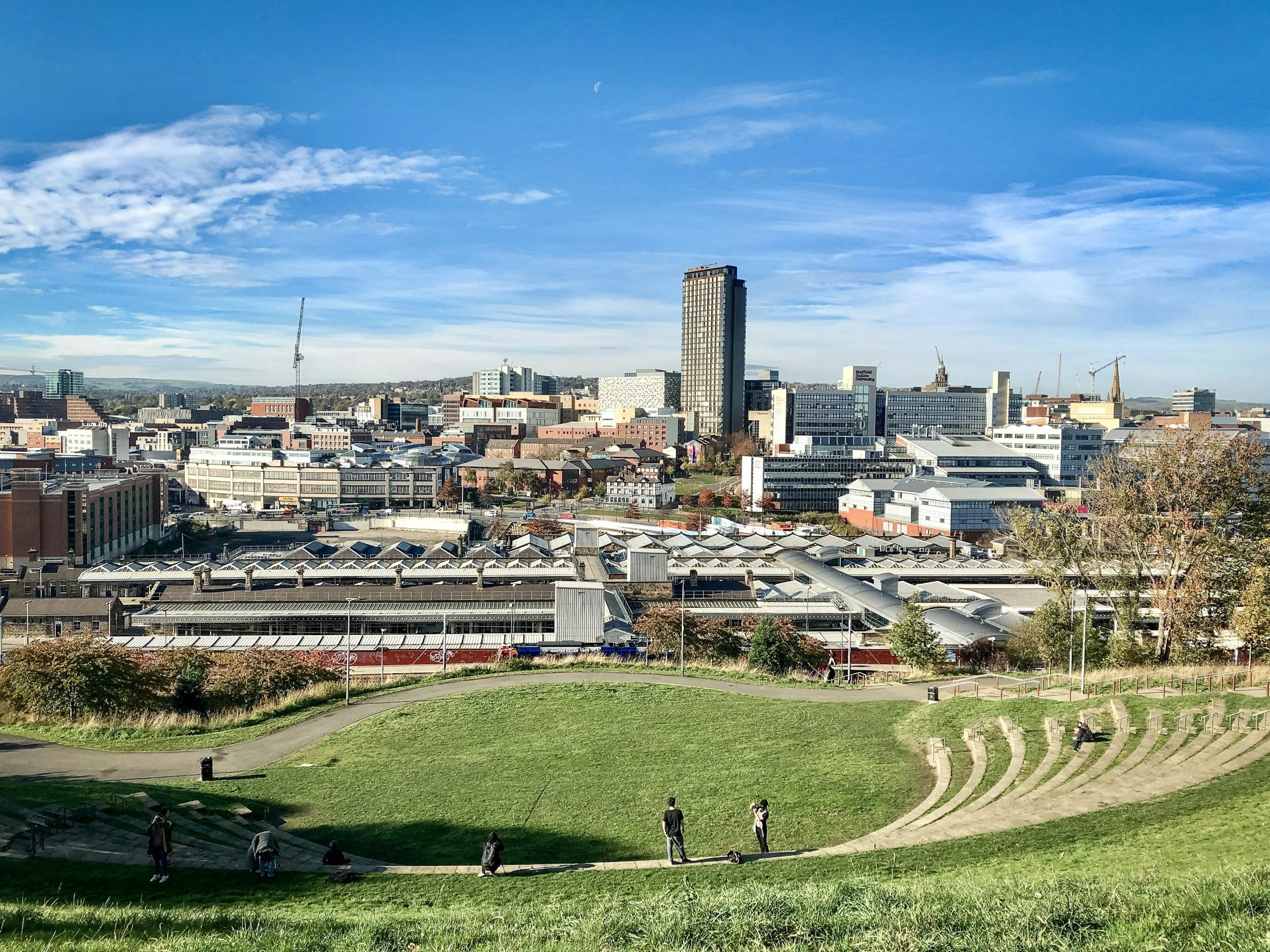 A Sheffield cityscape with a tall modern building under a bright blue sky with scattered clouds, seen from a park with grassy slopes and people walking or sitting.