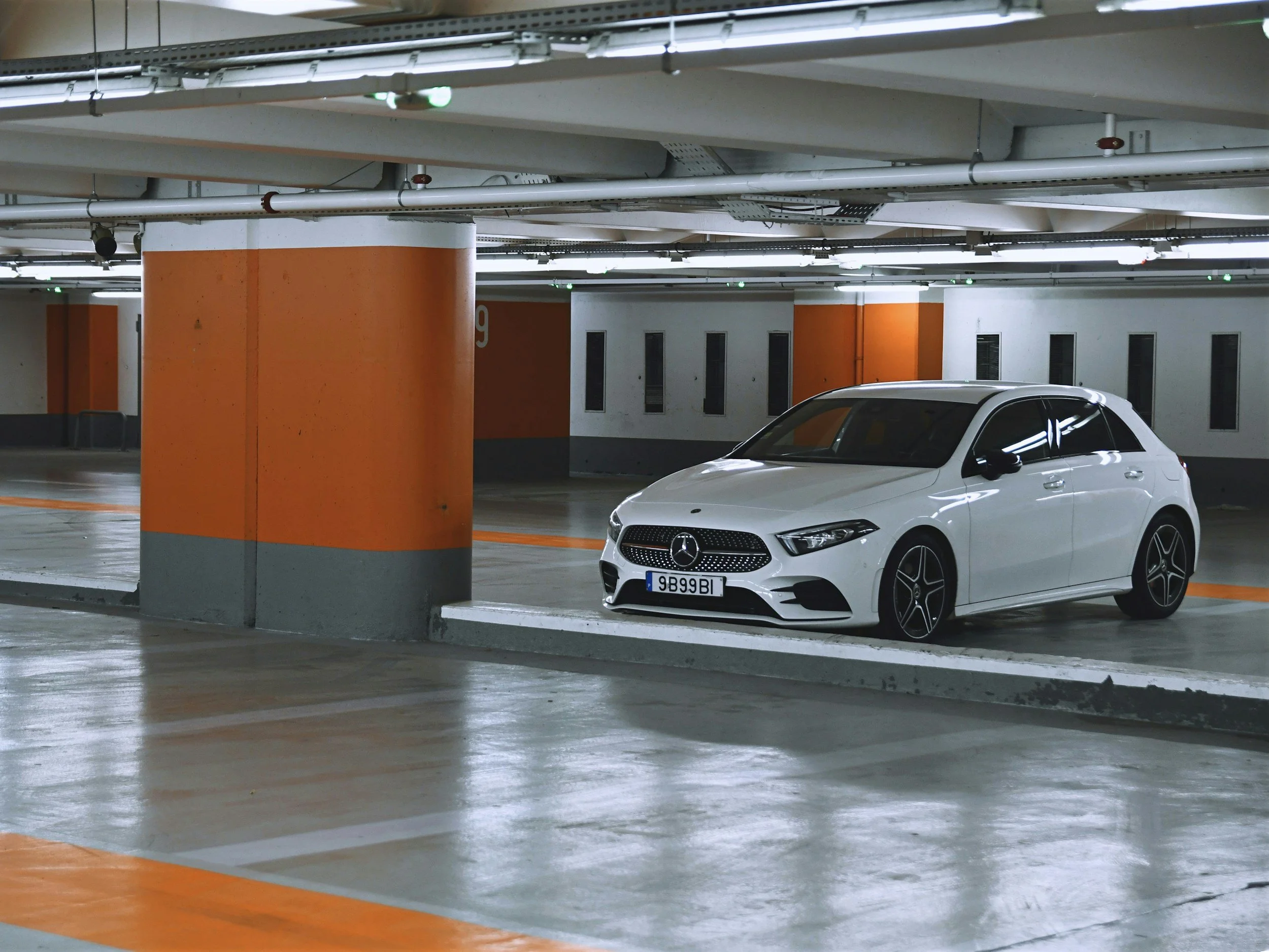 A white Mercedes-Benz car parked in an underground parking garage with orange and white painted pillars and a concrete ceiling with pipes and lighting.