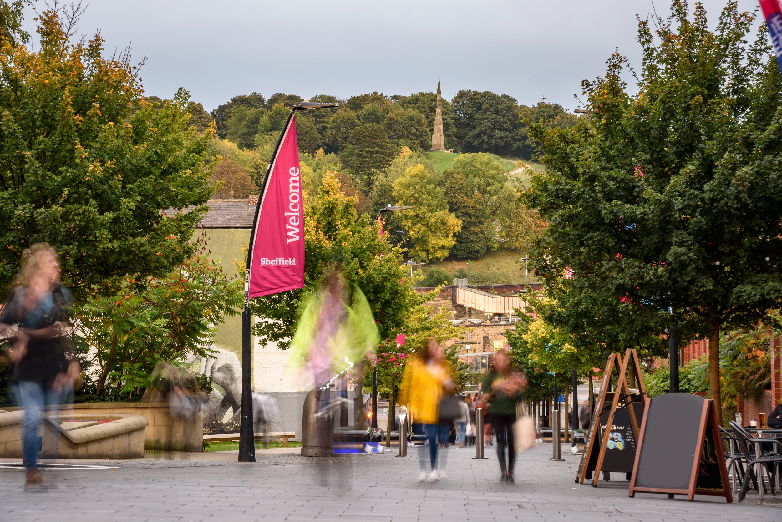 A street scene in Sheffield during a cloudy day with trees, a pink banner that says 'Welcome Sheffield', several blurred pedestrians, an outdoor seating area to the right, and a monument on a hill in the background.