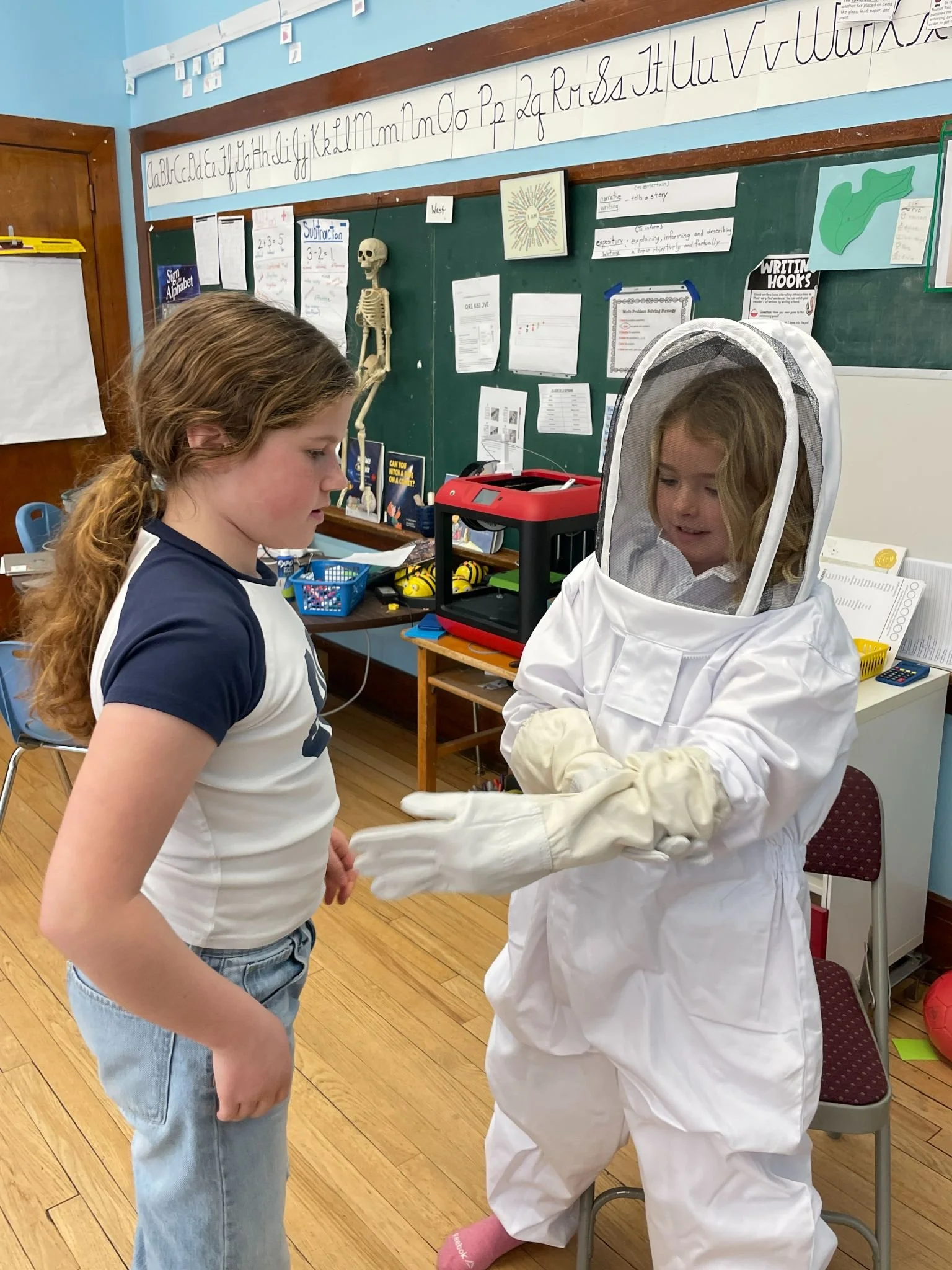 Two young girls in a classroom, one wearing a beekeeper suit, are talking. The classroom has a chalkboard, educational posters, and a skeleton model.