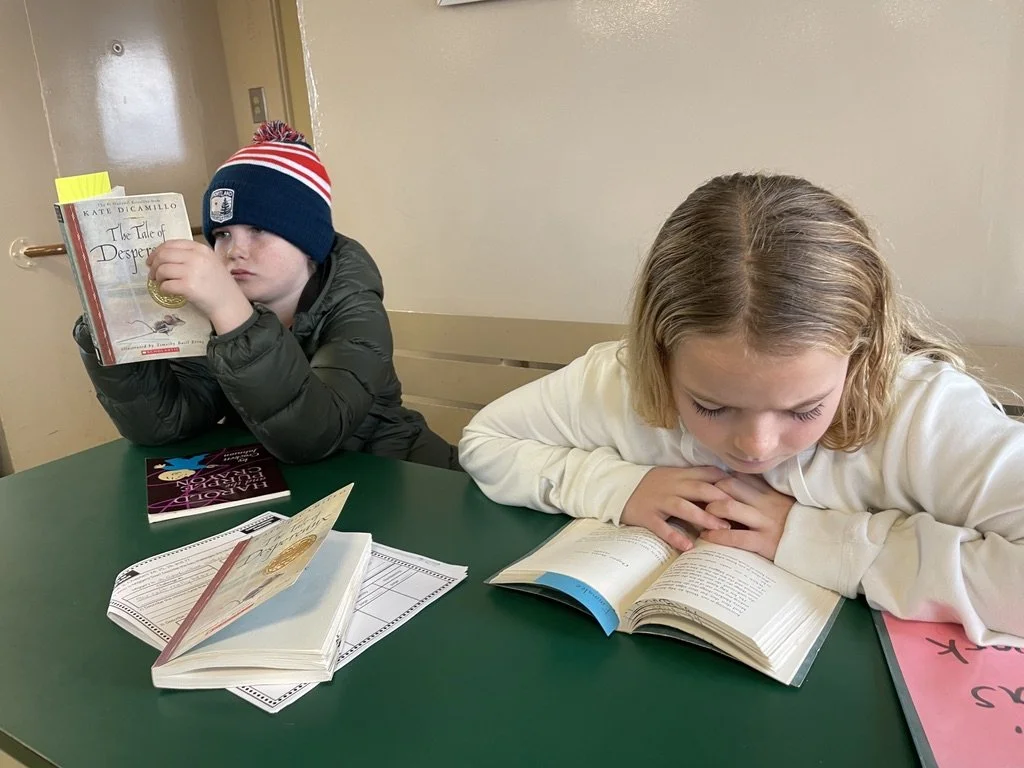 Two children sitting at a green table reading books. The girl on the right has blonde hair and is focused on her book, while the boy on the left wears a beanie and is looking at a different book.