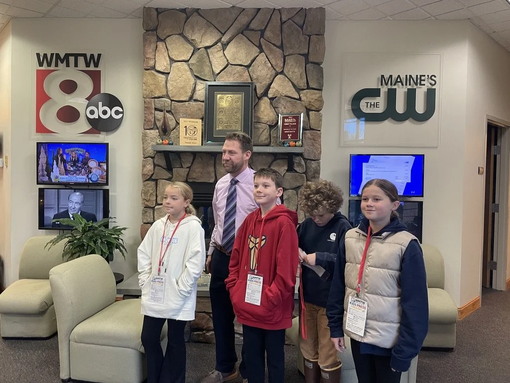 A group of four children and an adult standing in front of a stone fireplace inside a building. The children are wearing name tags and jackets, with some looking at the camera. The background displays TV screens and signs for WMTW ABC and Maine's CW.