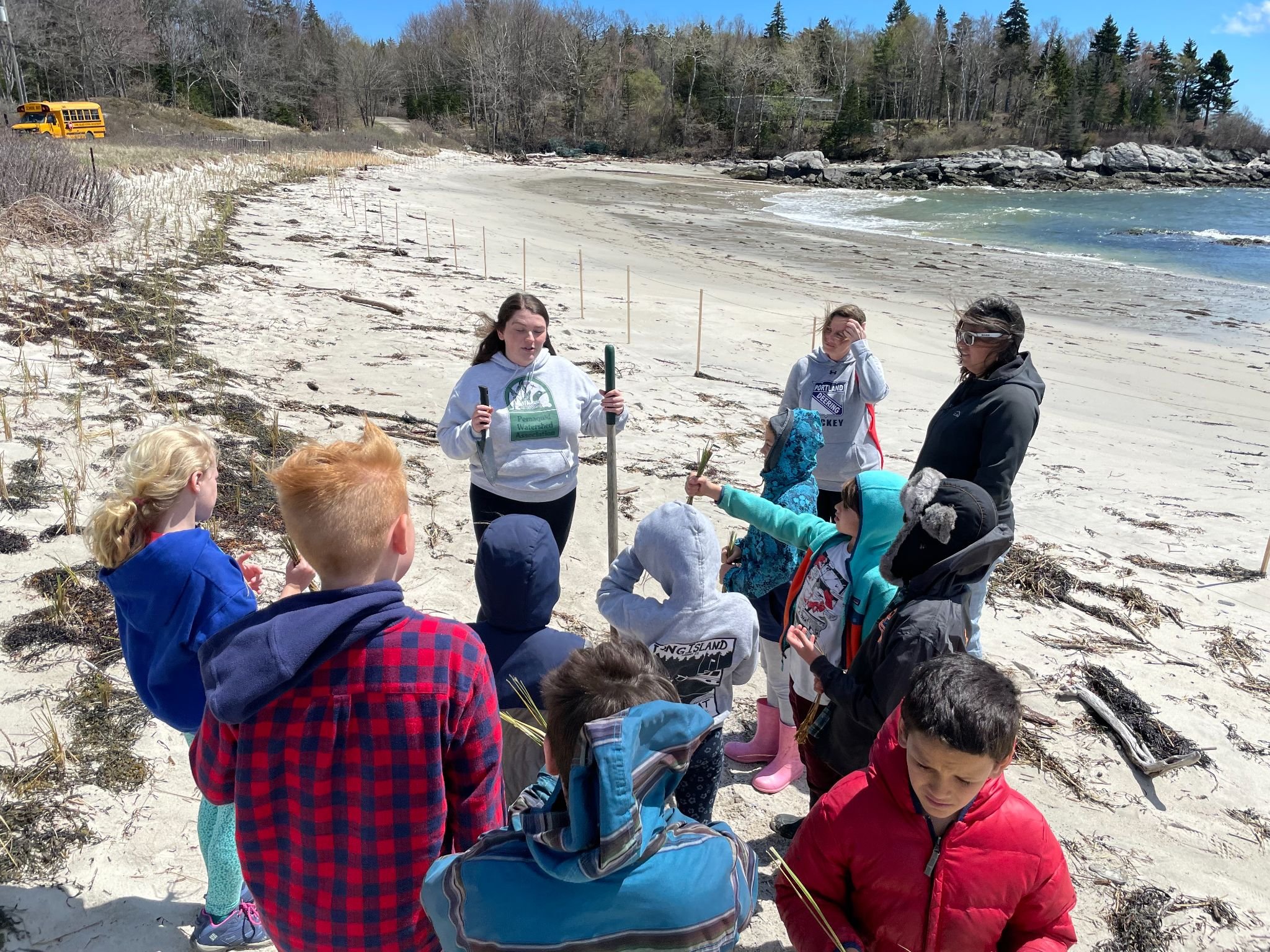 Group of children and adults on a sandy beach, listening to a woman in a gray hoodie describing something, with a forested area and a yellow school bus in the background on a clear day.