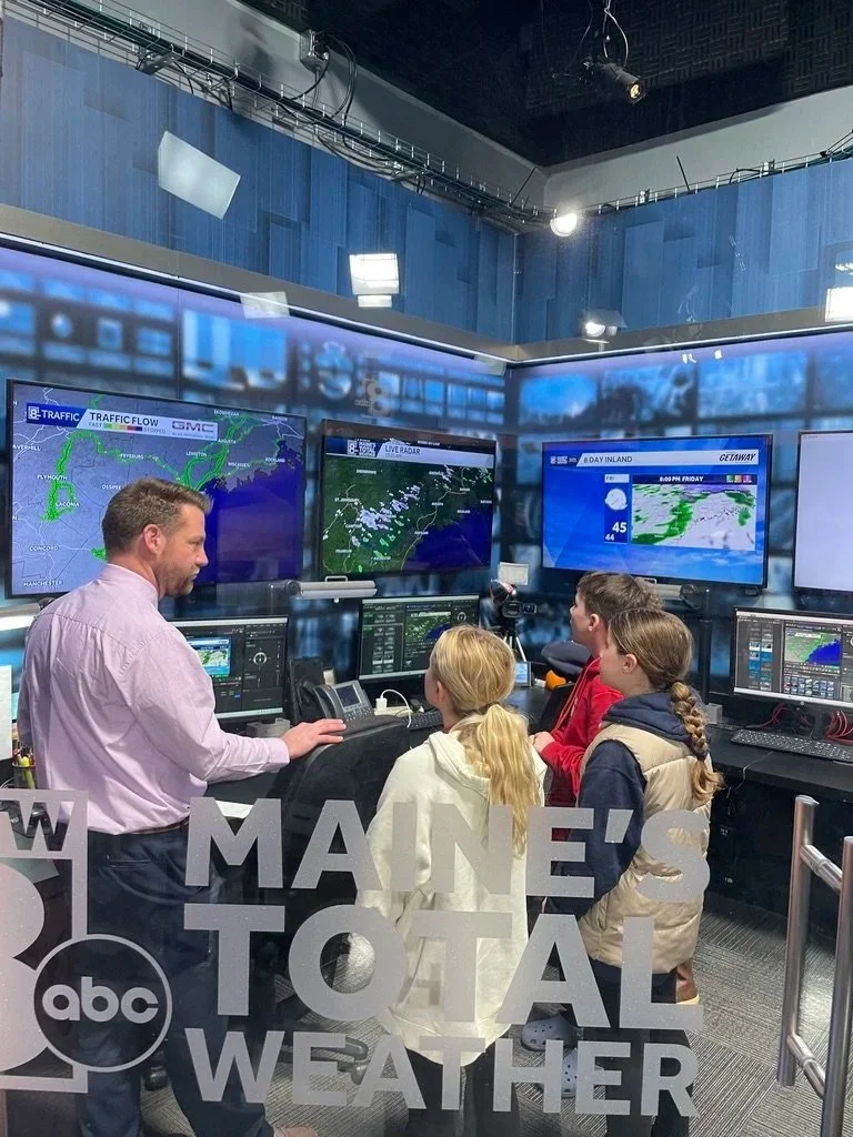 A man teaches children about weather monitoring inside a television station control room, with multiple screens displaying weather maps and traffic flow.