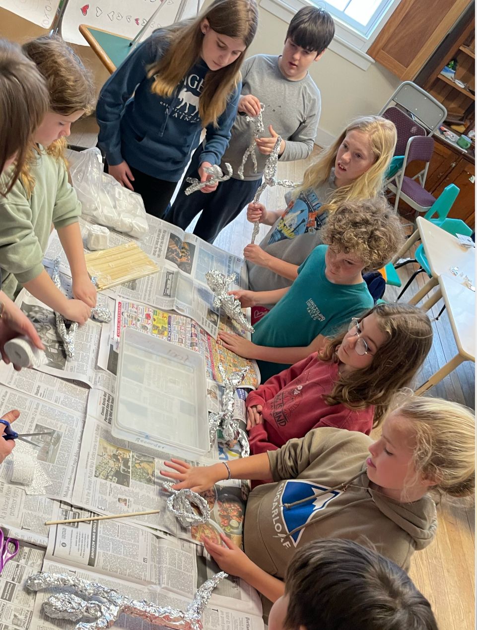 Children working together on a craft project with aluminum foil, some holding or shaping foil sculptures, at a table covered with newspapers.