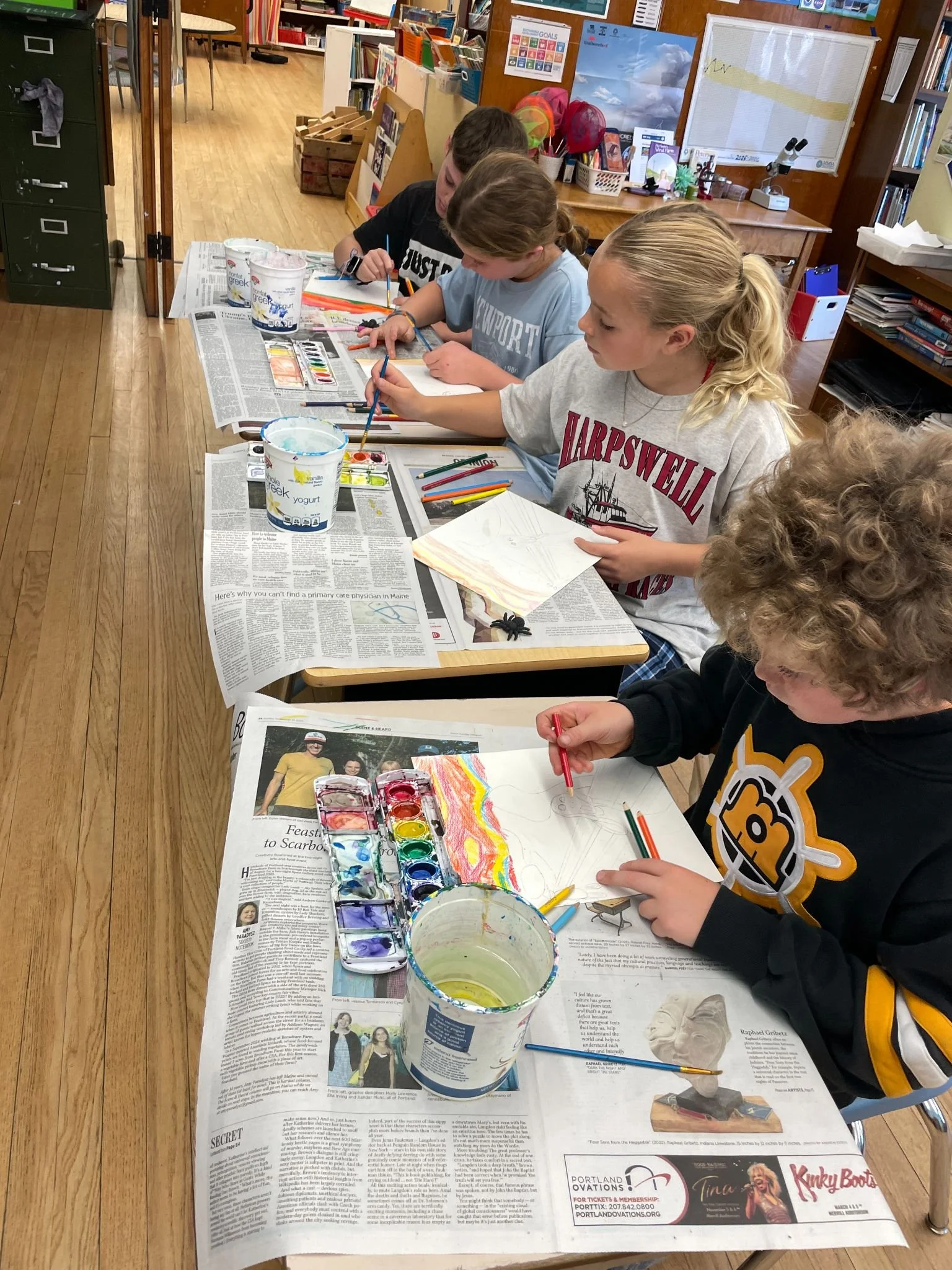 Children sitting at a table, painting with watercolor paints on paper, covered with newspapers to protect the surface, in a classroom or library setting.