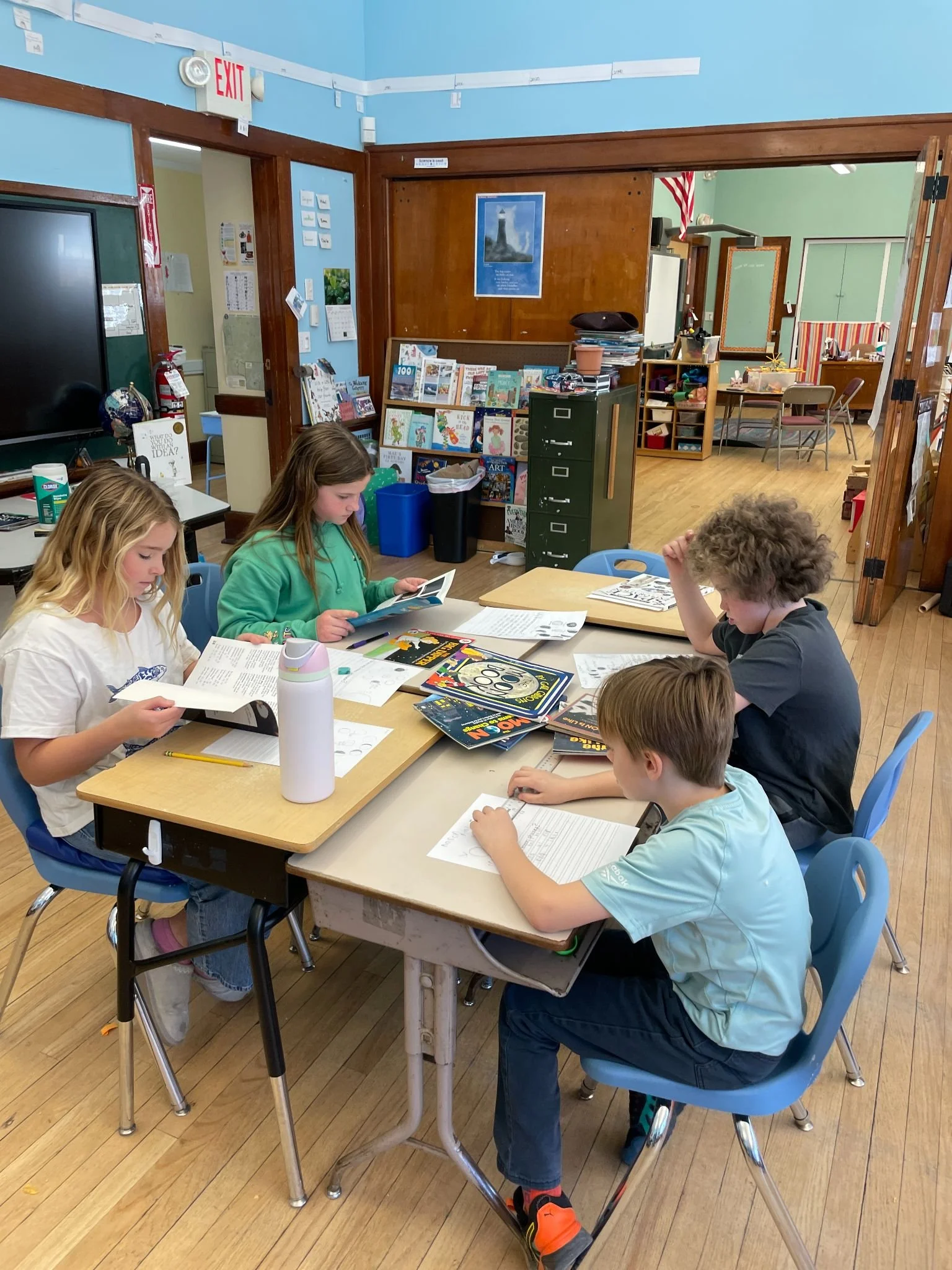 Children sitting at a table in a classroom, reading books and working on worksheets, with educational materials and furniture in the background.