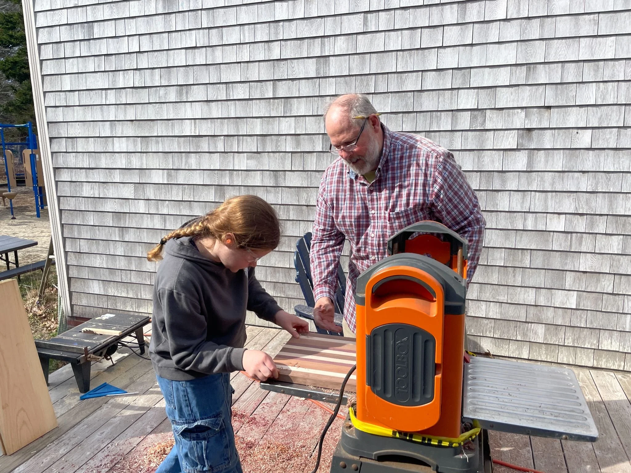 A man and a girl working with a saw on a wooden project on a workbench outside. The man is wearing a plaid shirt and glasses, the girl has a braid and is wearing a hoodie. There is a gray shingled wall in the background.