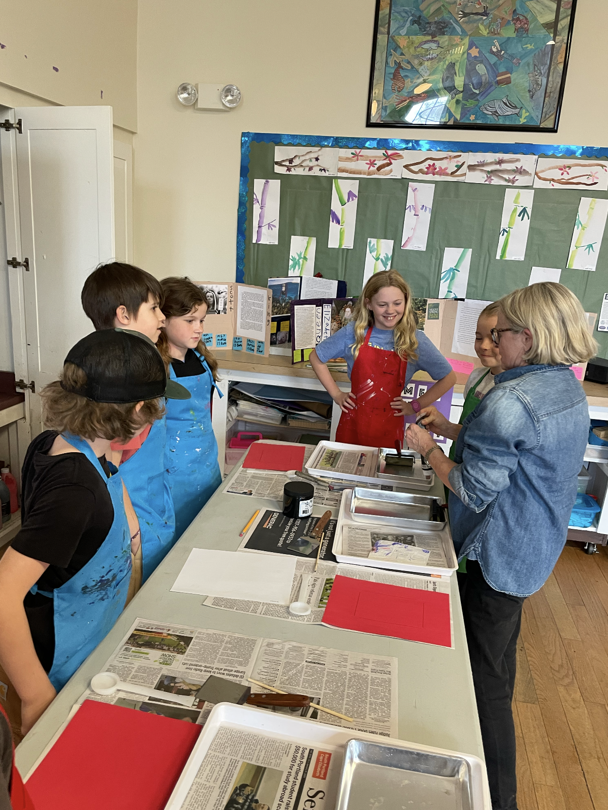 A group of children and an adult are gathered around a classroom table engaged in a science activity, with various supplies, newspapers, and red papers on the table. The background has a bulletin board and artwork displayed on the wall.