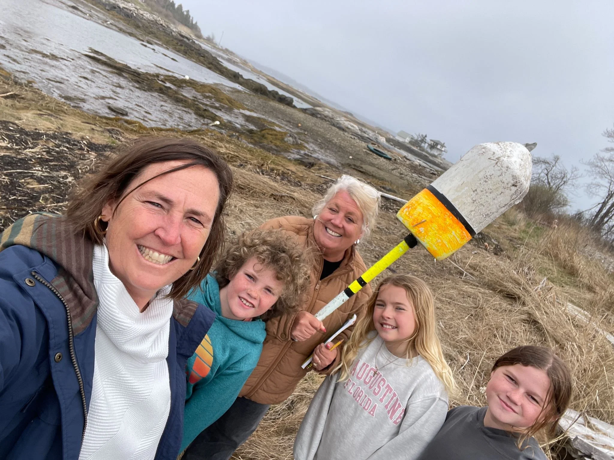 A group of smiling people, including children, standing outdoors on a cloudy day in a coastal area with muddy terrain and boats in the background, holding a yellow and black foam rocket.
