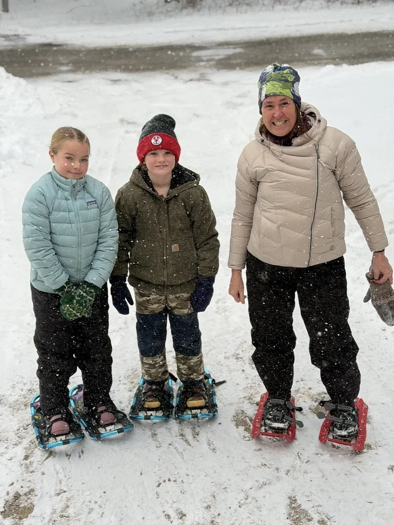 Three children and an adult standing on snow with snowshoes, wearing winter clothing and smiling.