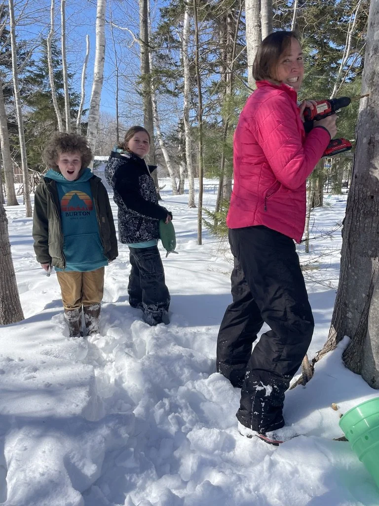 Three people in snowy woods, two children with snow boots and jackets, an adult woman with a drill in her hand, also in winter clothing, standing near trees for maple syrup.