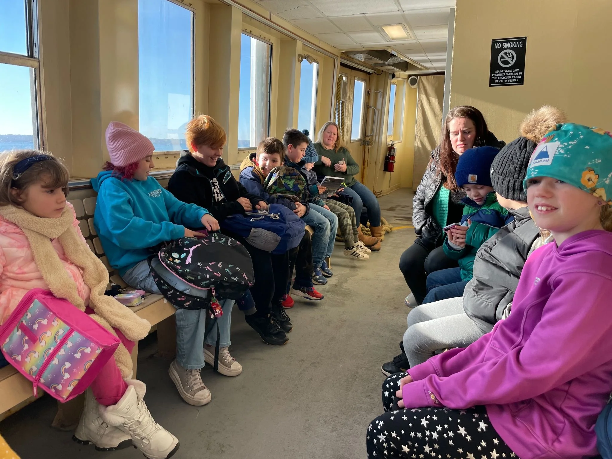 A group of children and two adults sitting on benches inside a ferry or boat, with large windows showing water outside. Some children are using electronic devices, and everyone is dressed warmly, indicating cool weather.