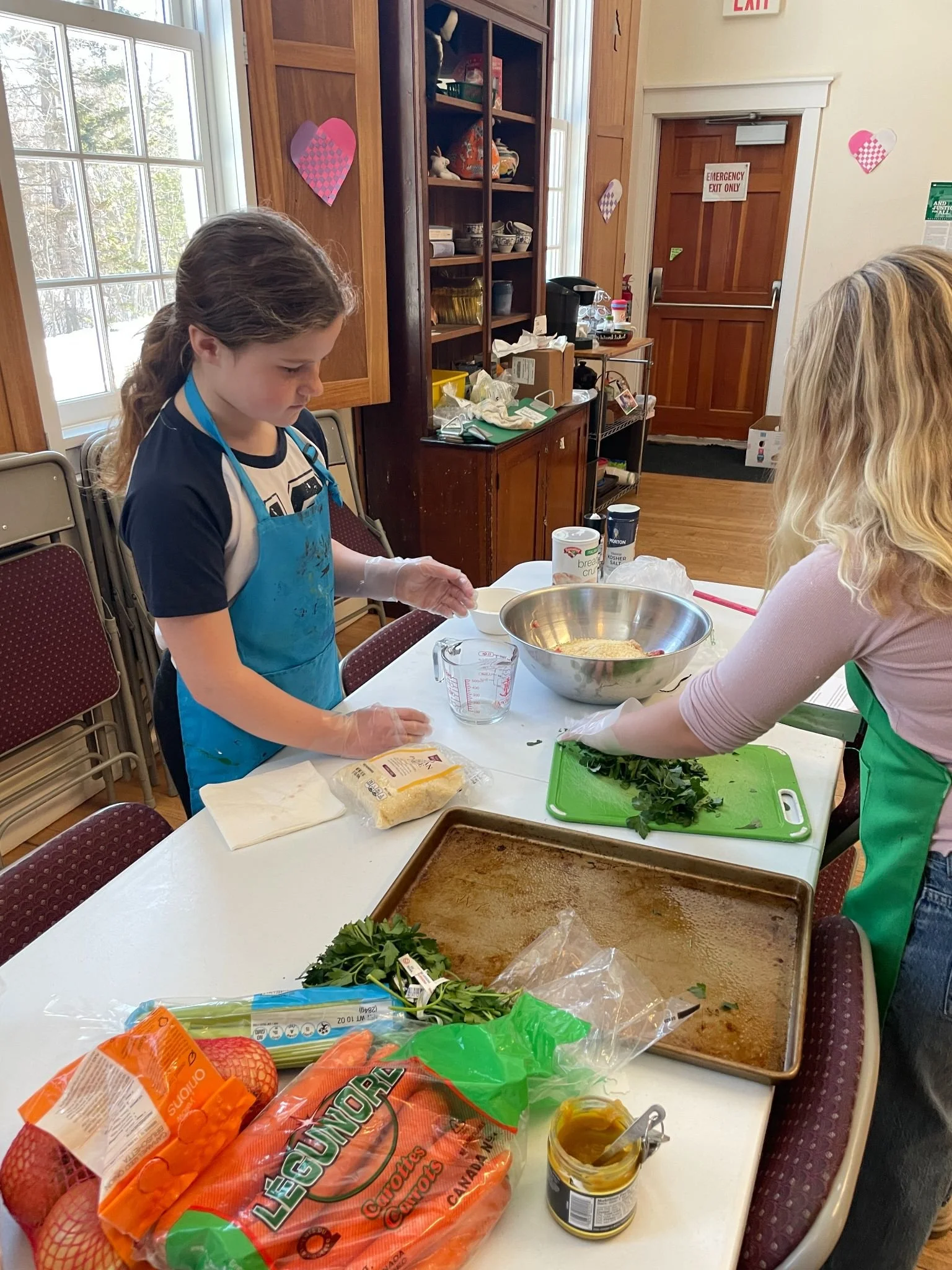 Two girls preparing food in a kitchen, one girl with long brown hair wearing a blue apron and the other with blonde hair wearing a green apron. They are working with various ingredients including a large bowl, chopped herbs, and packaged vegetables o