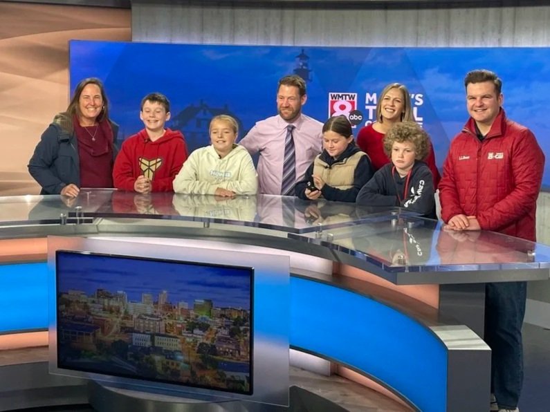 Group of eight people, including children and adults, standing behind a news desk in a television studio, with a backdrop featuring the WMTW 8 ABC logo and a cityscape on a monitor in front.