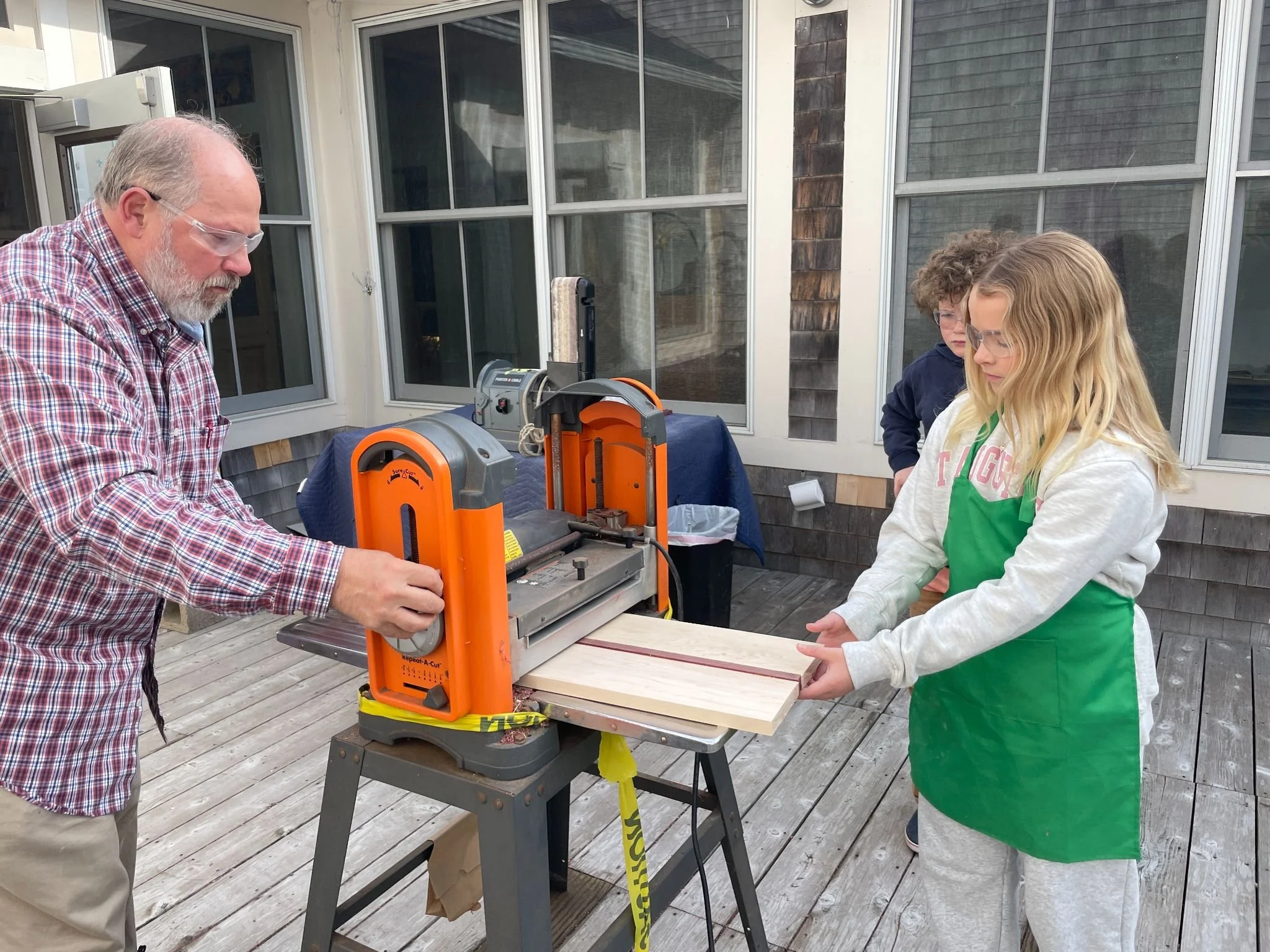Three people standing around an orange and black power saw on a work table outside on a wooden deck. One man with a beard and glasses is on the left, a young woman with long blonde hair and glasses is on the right, and a person with curly hair and gl