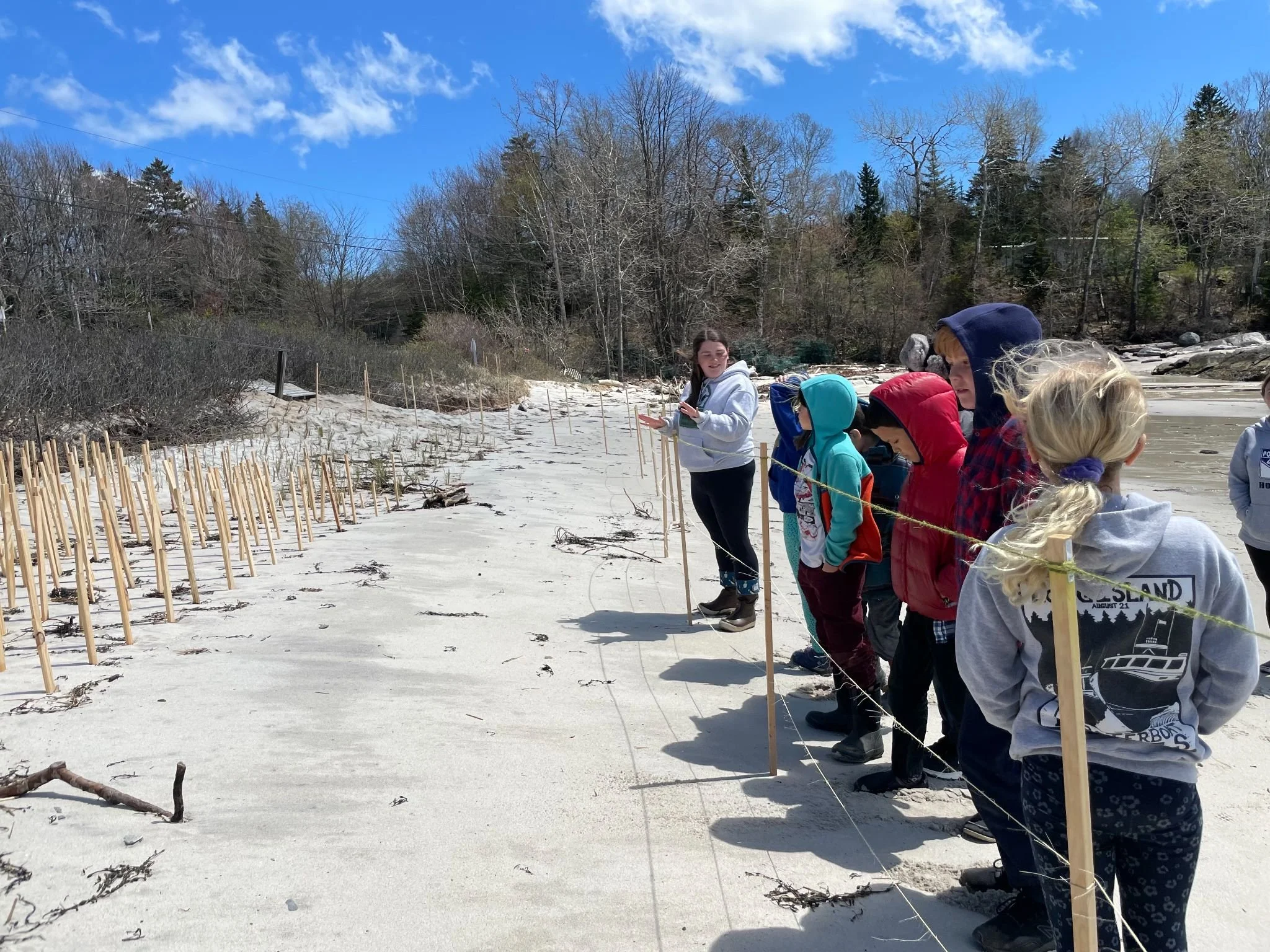 A group of children standing on a sandy beach, observing a line marked with small wooden sticks. The sky is blue with some clouds, and trees line the background. The children are dressed in jackets and hoodies.