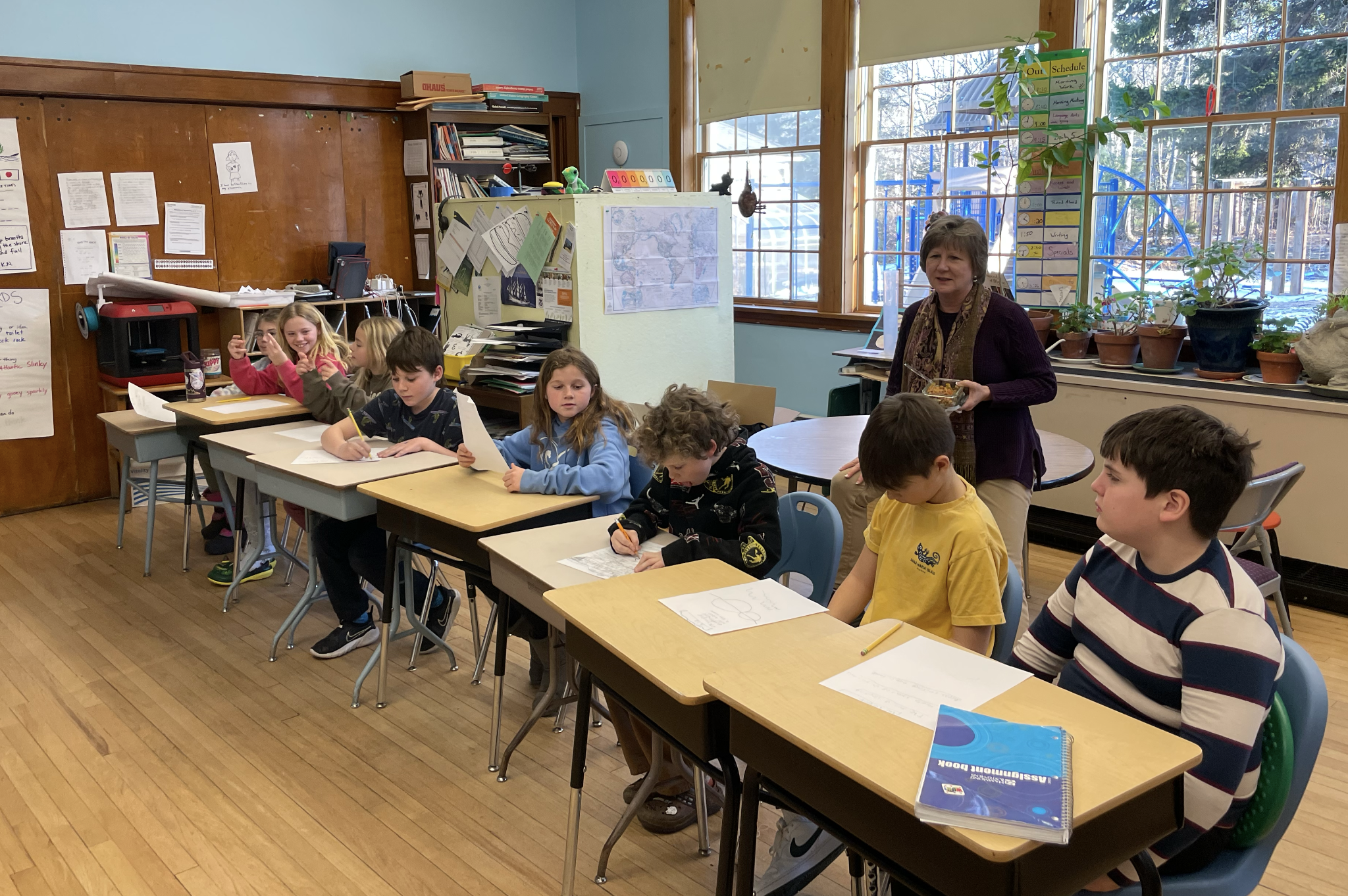 A classroom with students sitting at desks, a teacher standing with a bowl, large windows, and classroom decorations.