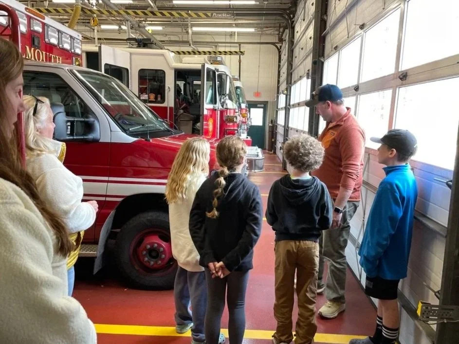 A group of children and a fire station staff member inside a fire station garage, standing near red fire trucks. The children are listening to the staff member, who appears to be explaining something about the fire trucks.