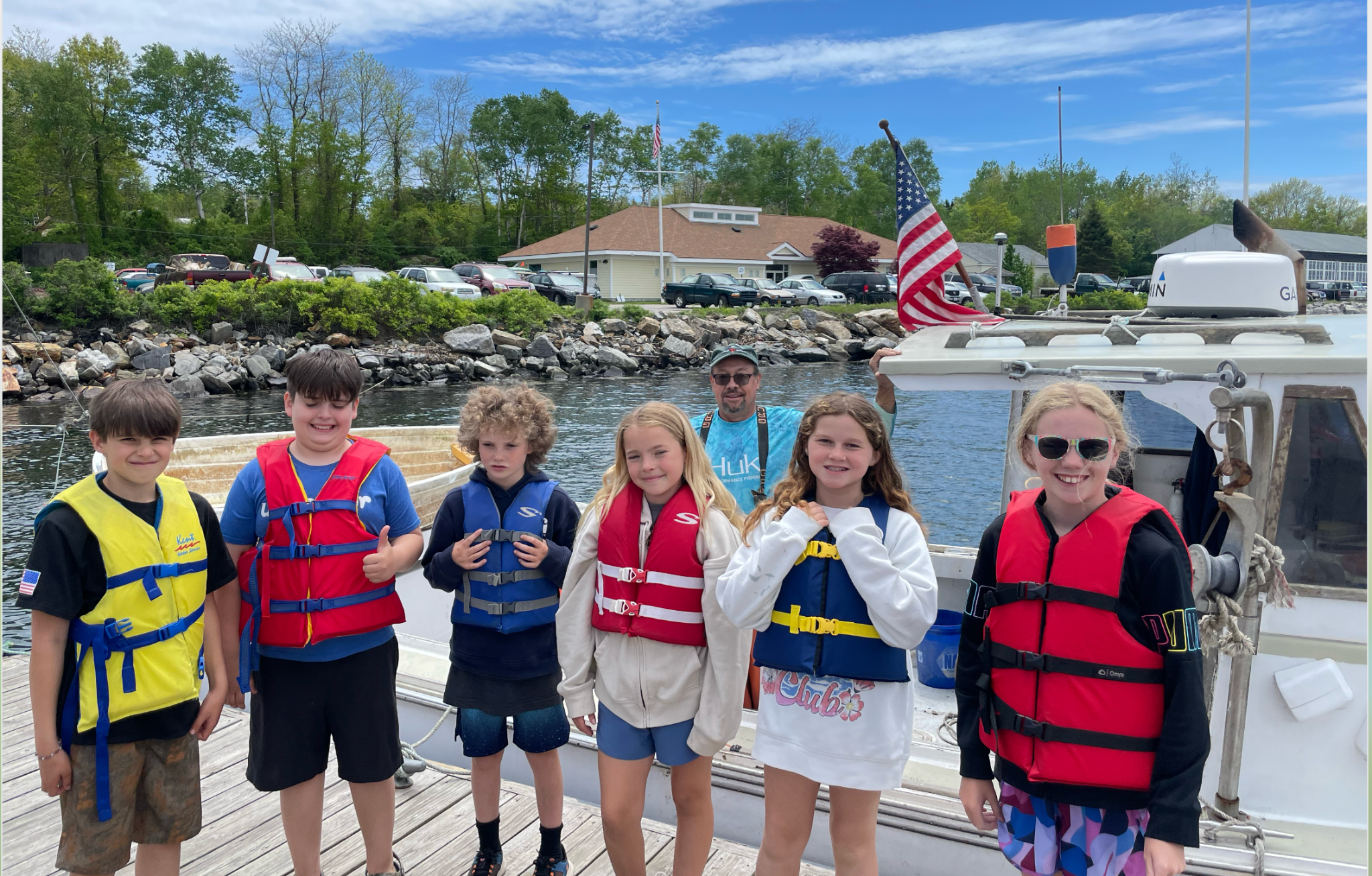 Group of six children and one adult on a boat docked near rocky shoreline, wearing life jackets, smiling, with trees, parked cars, and a building in the background on a partly cloudy day.