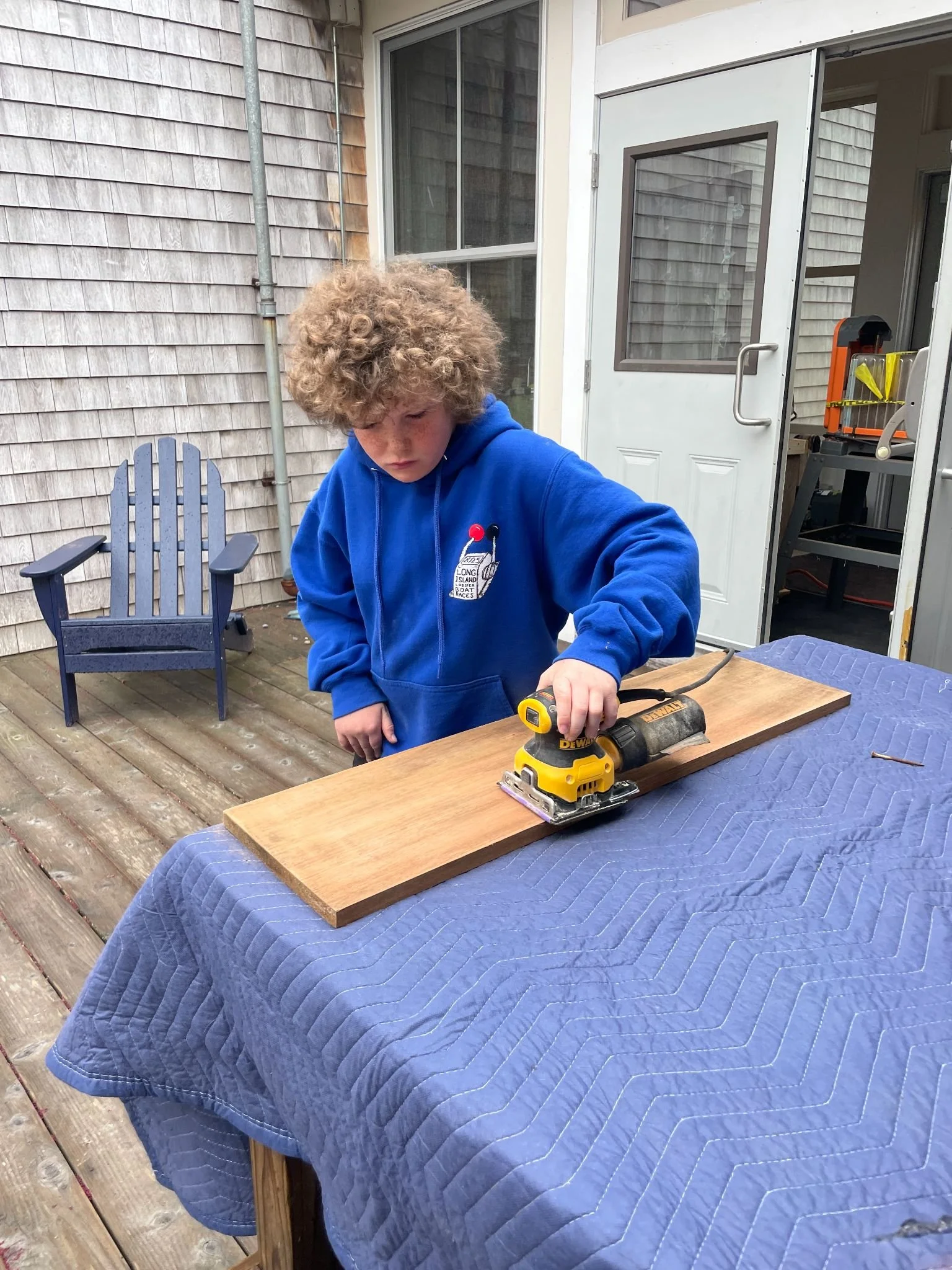 A young person with curly hair wearing a blue hoodie is using a power sander on a wooden piece outdoors on a table covered with a blue quilted cloth. There is a wooden deck, a chair, and a building with shingle siding in the background.