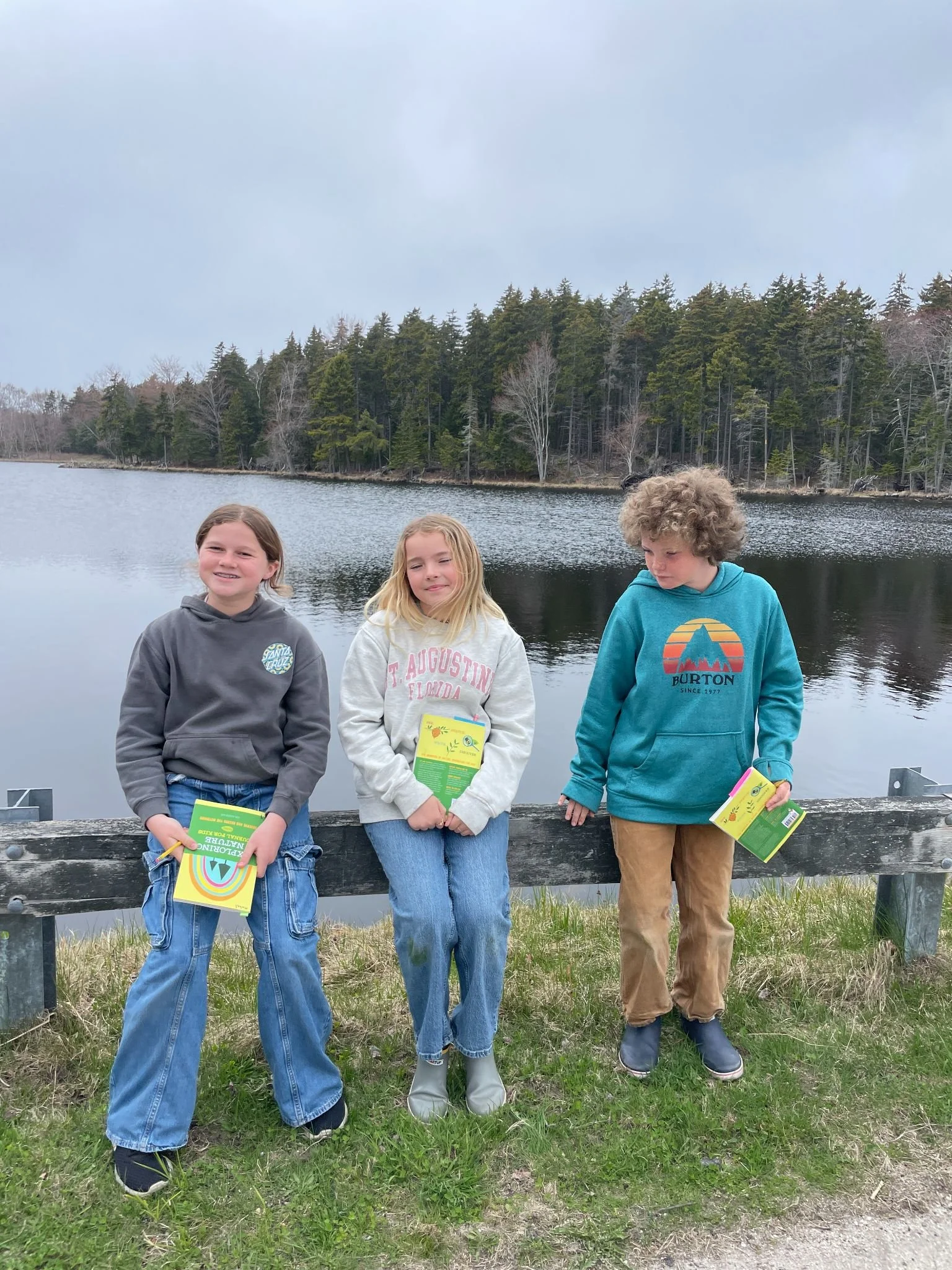 Three children standing by a lake with trees in the background, holding colorful brochures.
