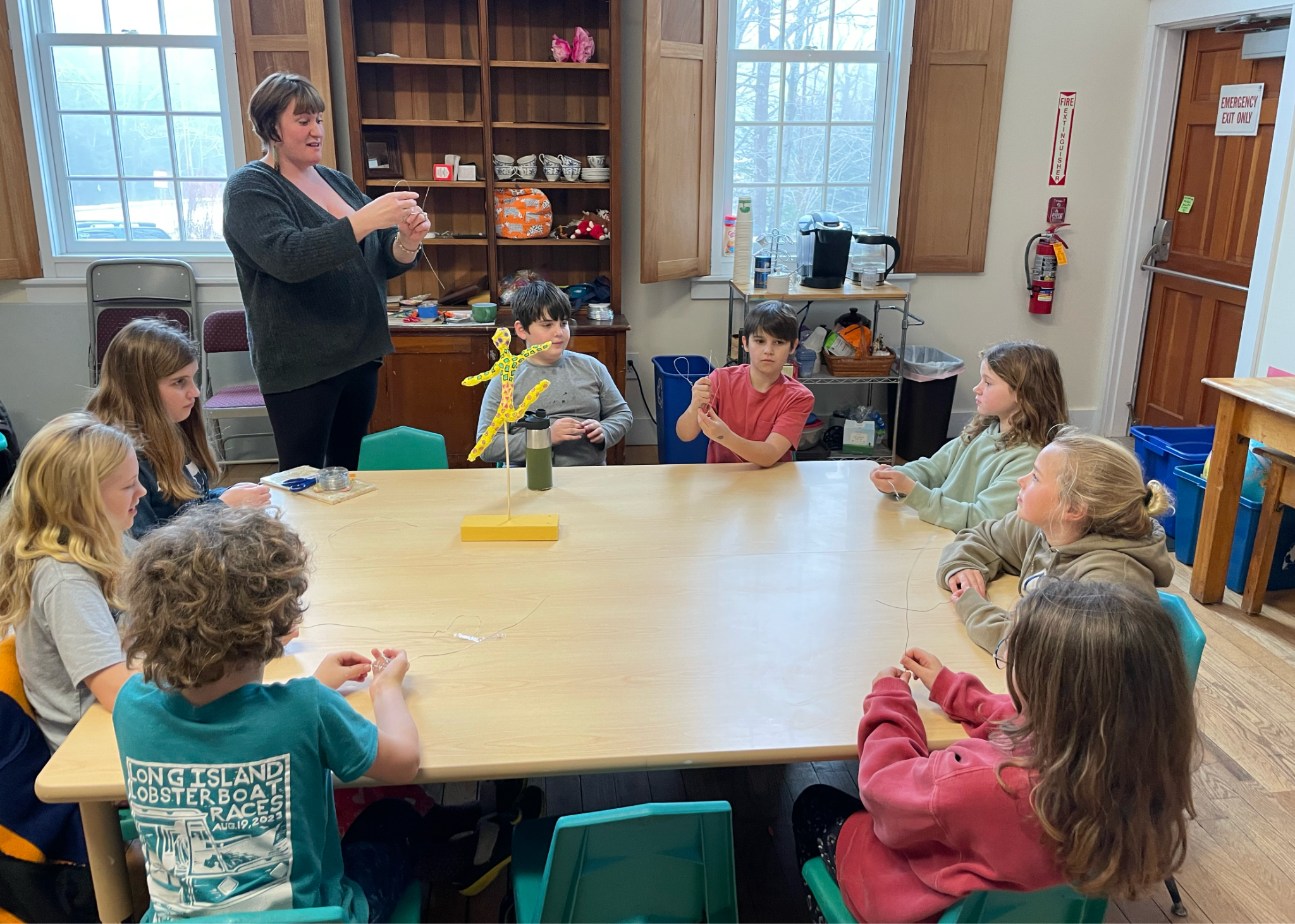A group of children sitting around a large wooden table in a room, with a woman standing and holding a string. The children are watching her and some have their hands raised or are holding objects. The room has windows, wooden cabinets, and shelves w