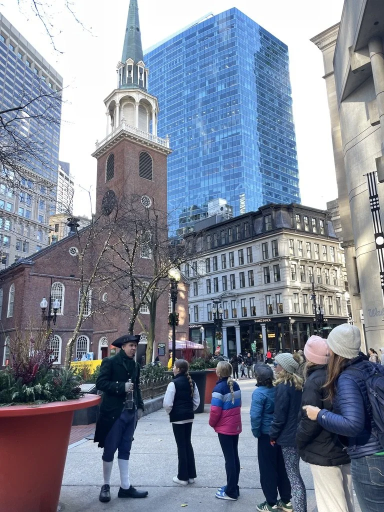 Tour guide dressed in historical costume talking to a group of children in a city square, with a brick church and modern glass buildings in the background.