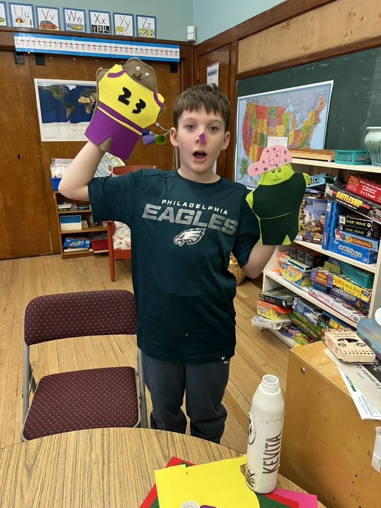 A boy wearing a Philadelphia Eagles shirt holding two puppets, one on each hand, in a classroom setting with educational posters, a map of the United States, and shelves with books and puzzles.