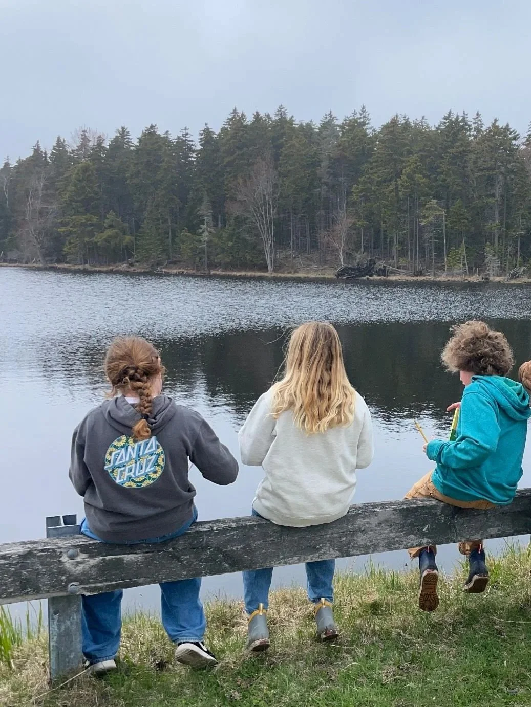 Three children sit on a wooden bench facing a calm lake with a forested shoreline in the background. They are wearing casual clothes, with one girl wearing a Santa Cruz hoodie, and one holding a pair of yellow chopsticks or sticks.