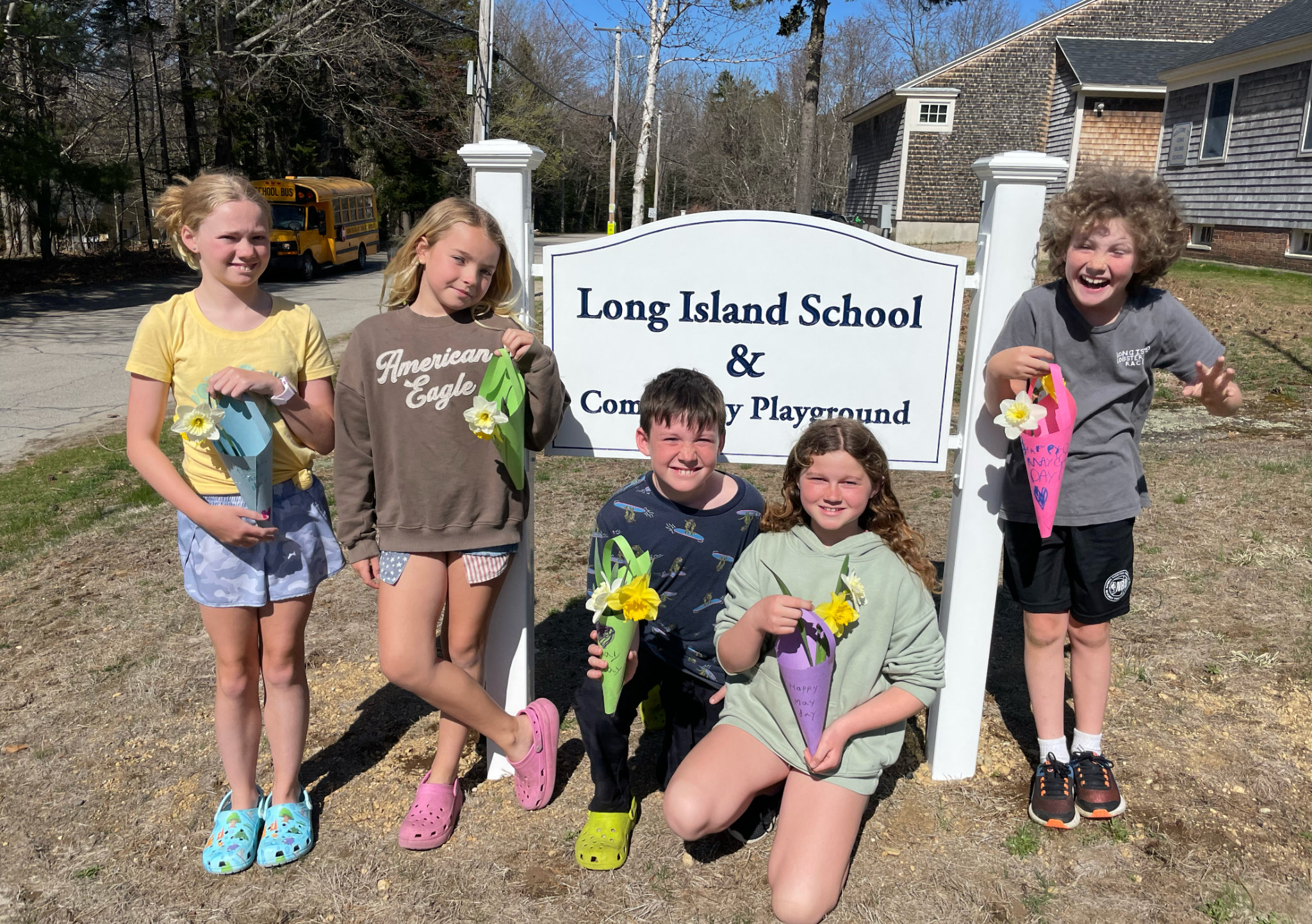 Group of six children standing outdoors near a sign that reads 'Long Island School & Community Playground'. The children are holding decorated flower cones and smiling, with a school bus and houses in the background on a sunny day.