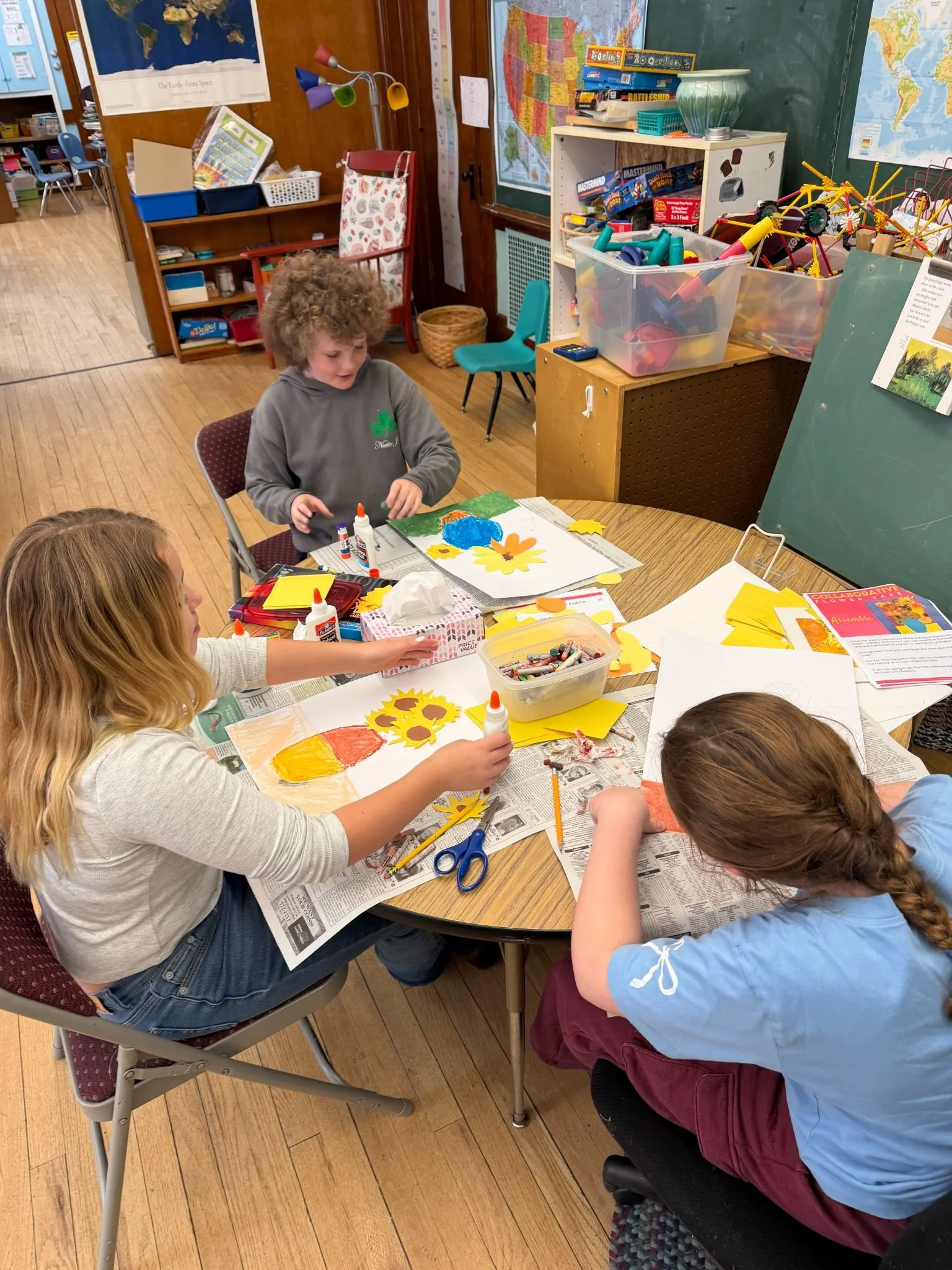 Children sitting around a table engaging in arts and crafts activities, with various craft supplies such as glue, scissors, and paper on the table. A classroom setting with educational posters and maps on the walls.